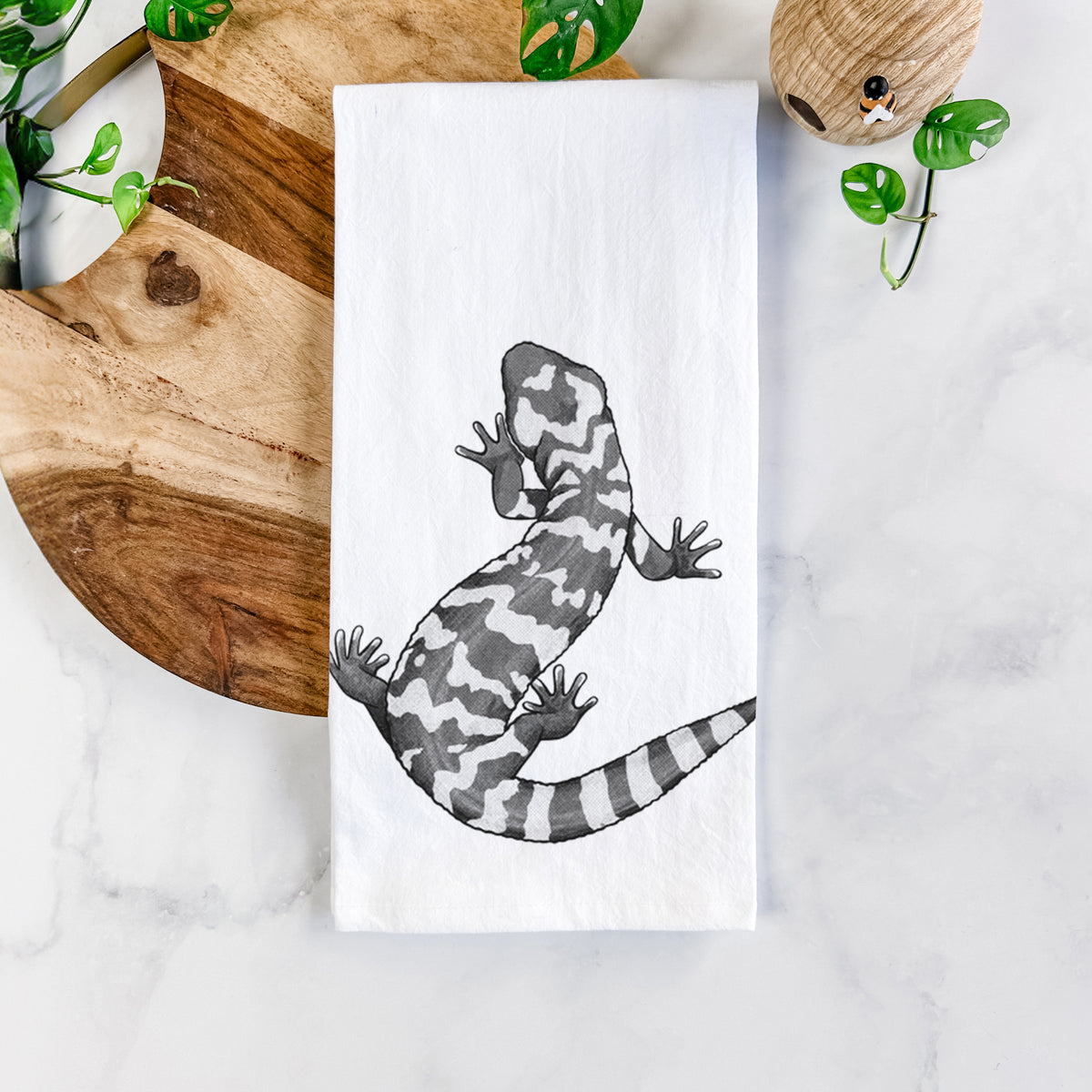 White cotton tea towel printed with a black ink illustration of a Gila monster with bold patterned skin, on a marble counter beside a round wooden cutting board and greenery.