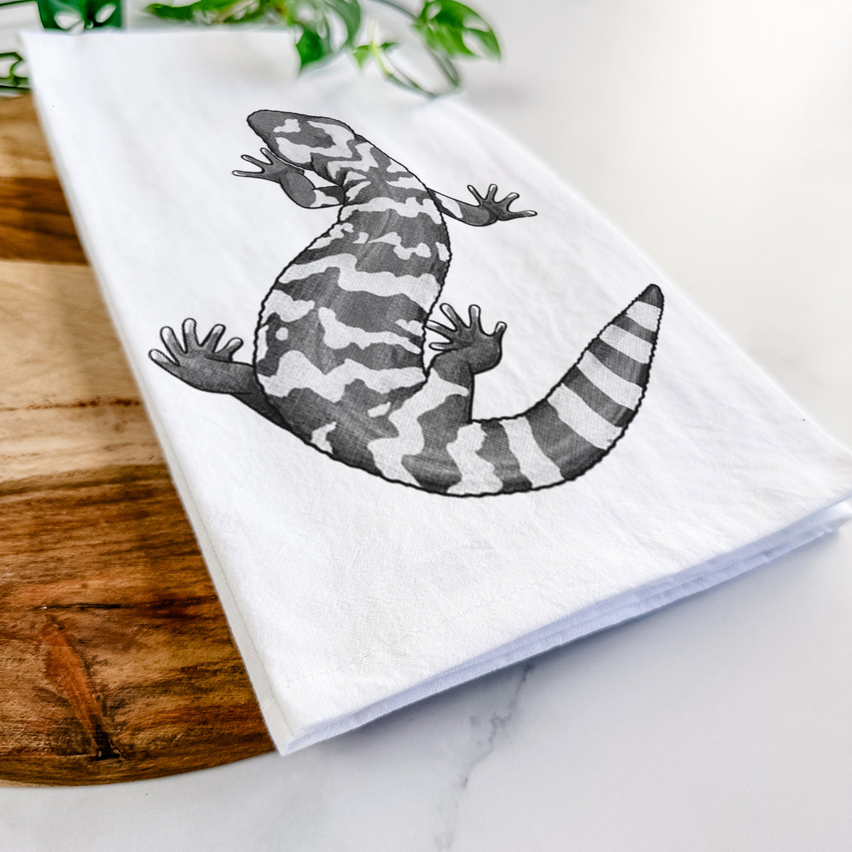 Close-up of a folded white tea towel on a wooden board, showing a black ink illustration of a Gila monster with bold patterned skin.