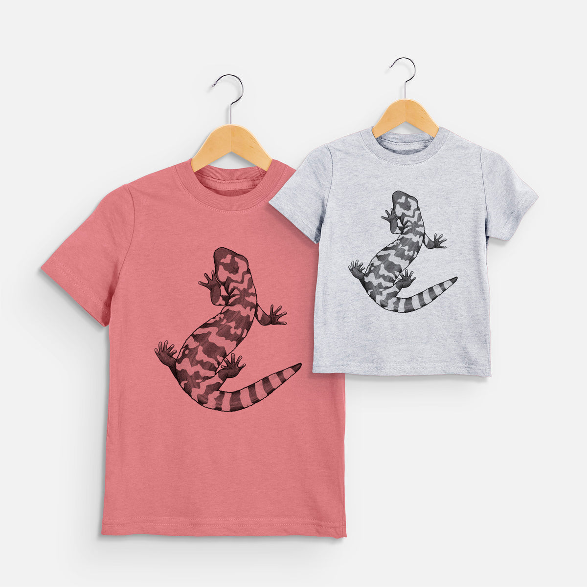 Two youth t-shirts in different colors on hangers, printed with a black ink illustration of a Gila monster with bold patterned skin.