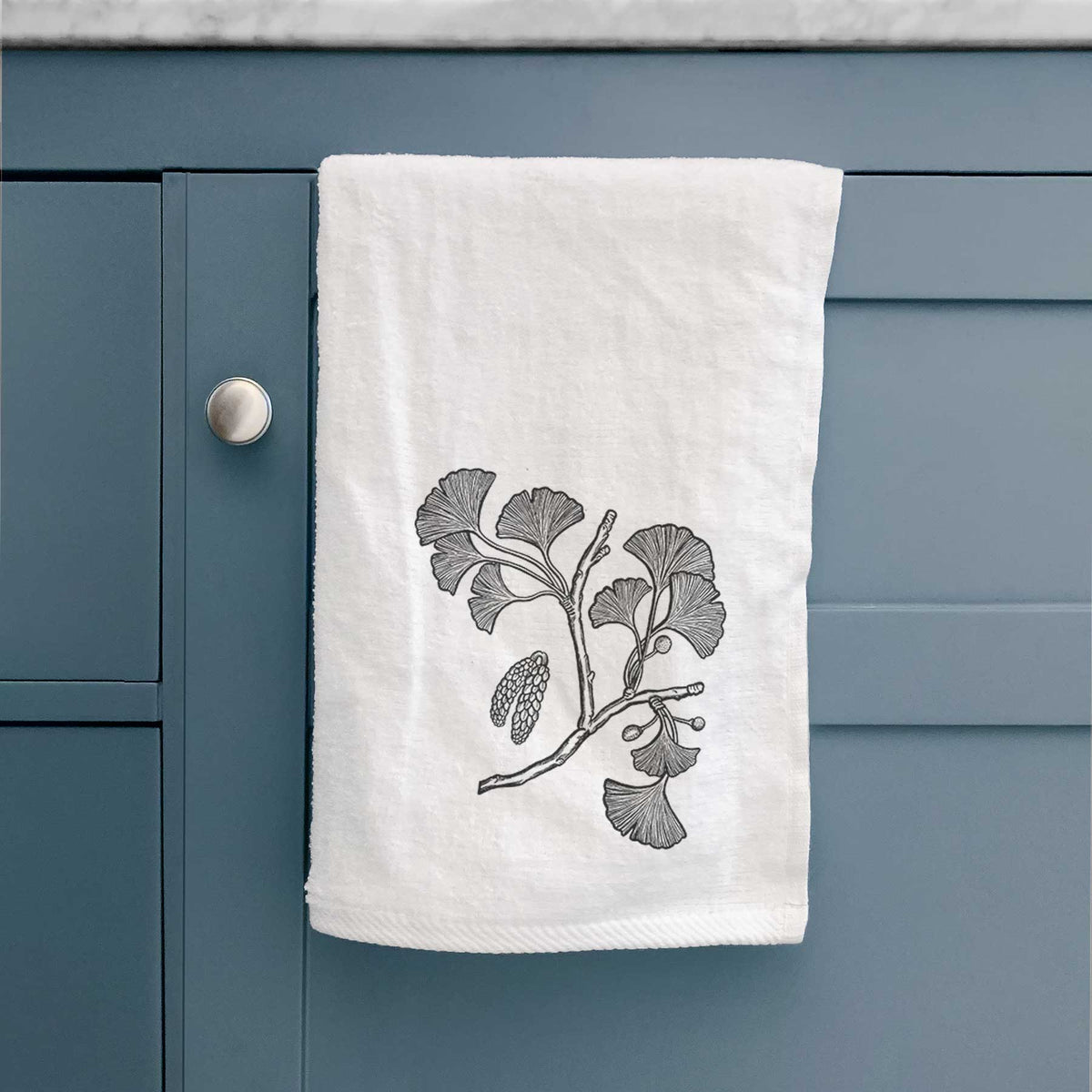 White hand towel printed with a black ink illustration of a ginkgo branch with fan-shaped leaves, hanging on a cabinet door.