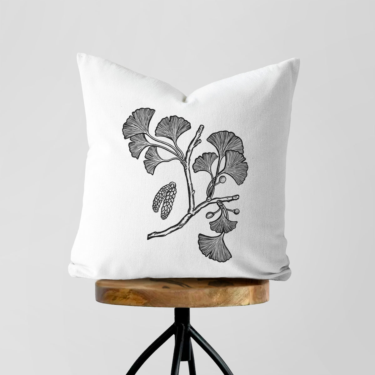 White pillow cover printed with a black ink illustration of a ginkgo branch with fan-shaped leaves, on a wooden stool.
