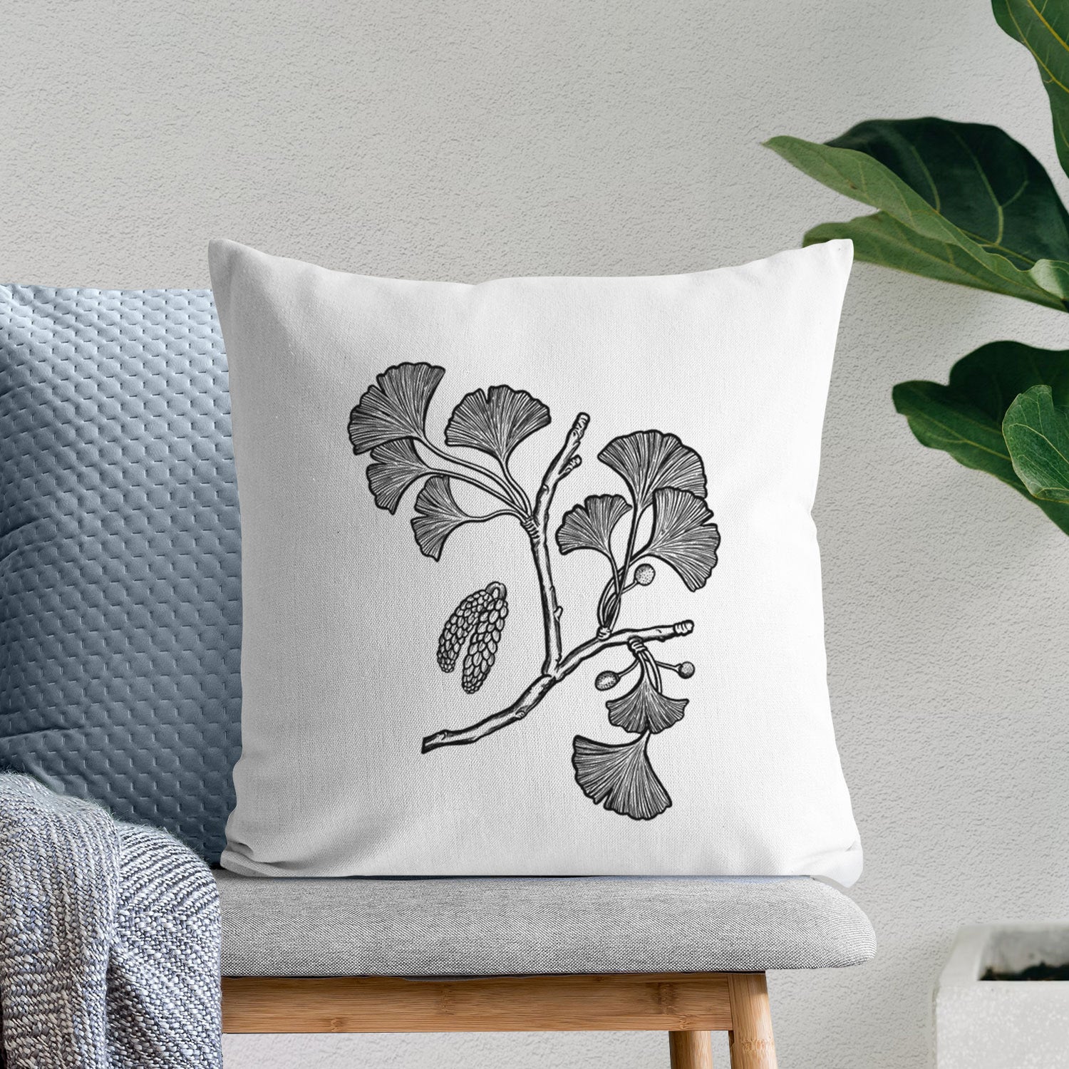 White pillow cover printed with a black ink illustration of a ginkgo branch with fan-shaped leaves, on a bench beside plants.