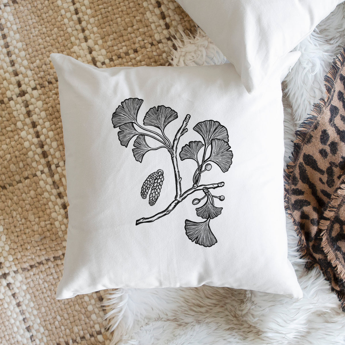 White pillow cover printed with a black ink illustration of a ginkgo branch with fan-shaped leaves, on a woven chair.