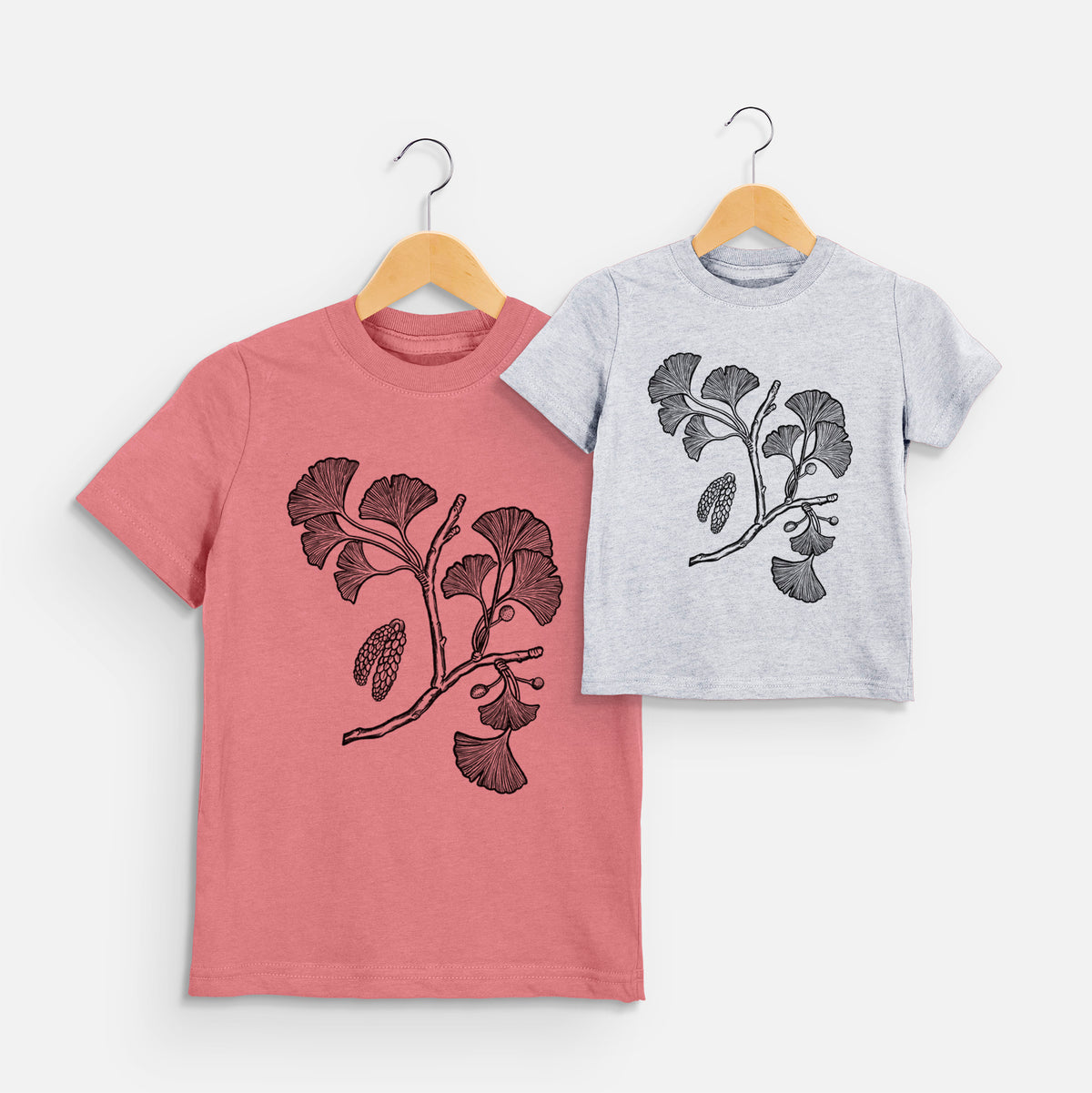Two youth t-shirts in different colors on hangers, printed with a black ink illustration of a ginkgo branch with fan-shaped leaves.