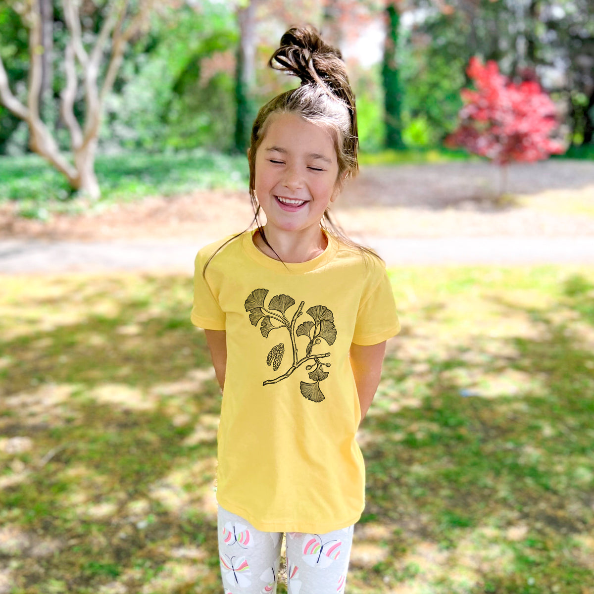 Smiling girl wearing a youth t-shirt printed with a black ink illustration of a ginkgo branch with fan-shaped leaves, outdoors.