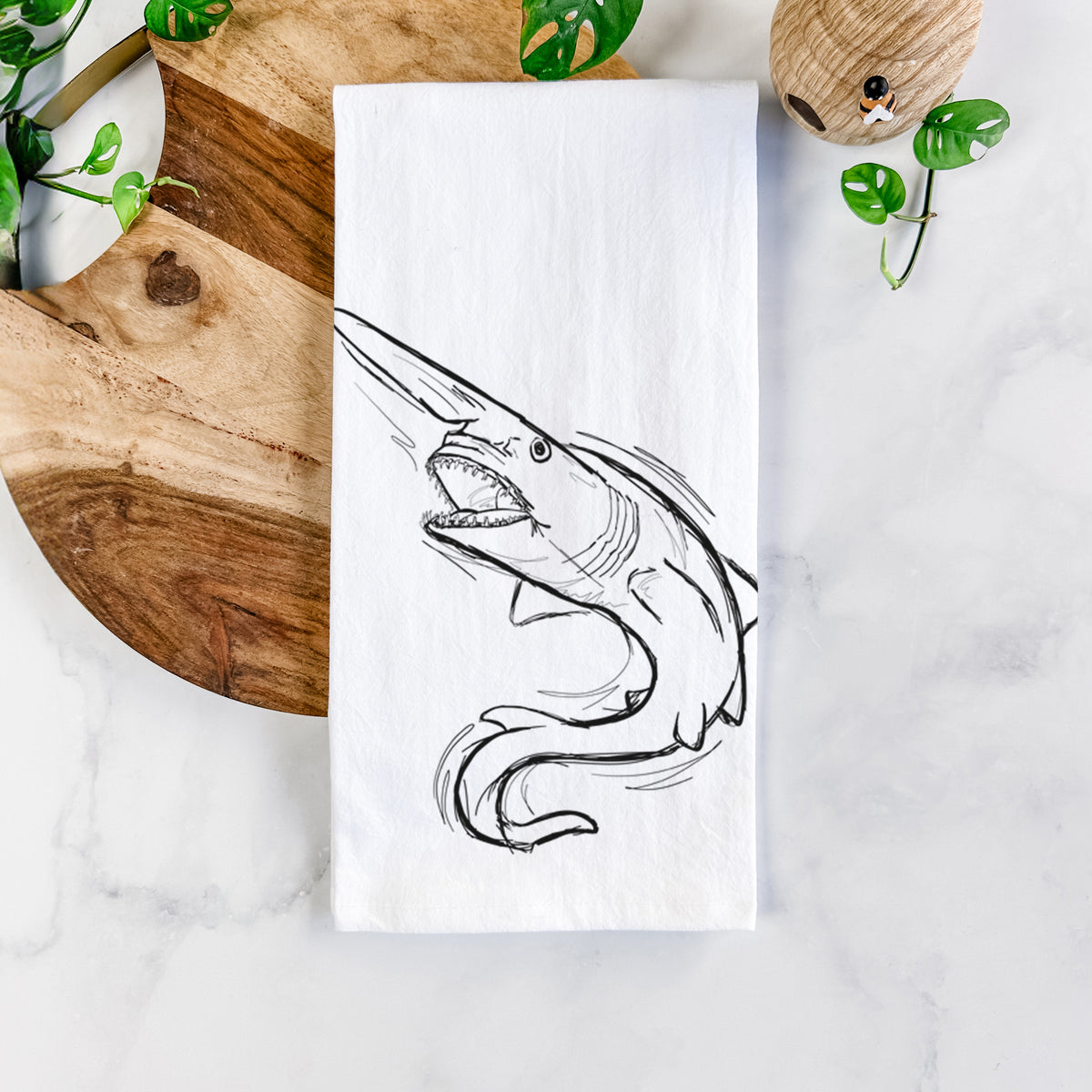 White cotton tea towel printed with a black ink illustration of a goblin shark with its long snout, swimming in a curve, on a marble counter beside a round wooden cutting board and greenery.
