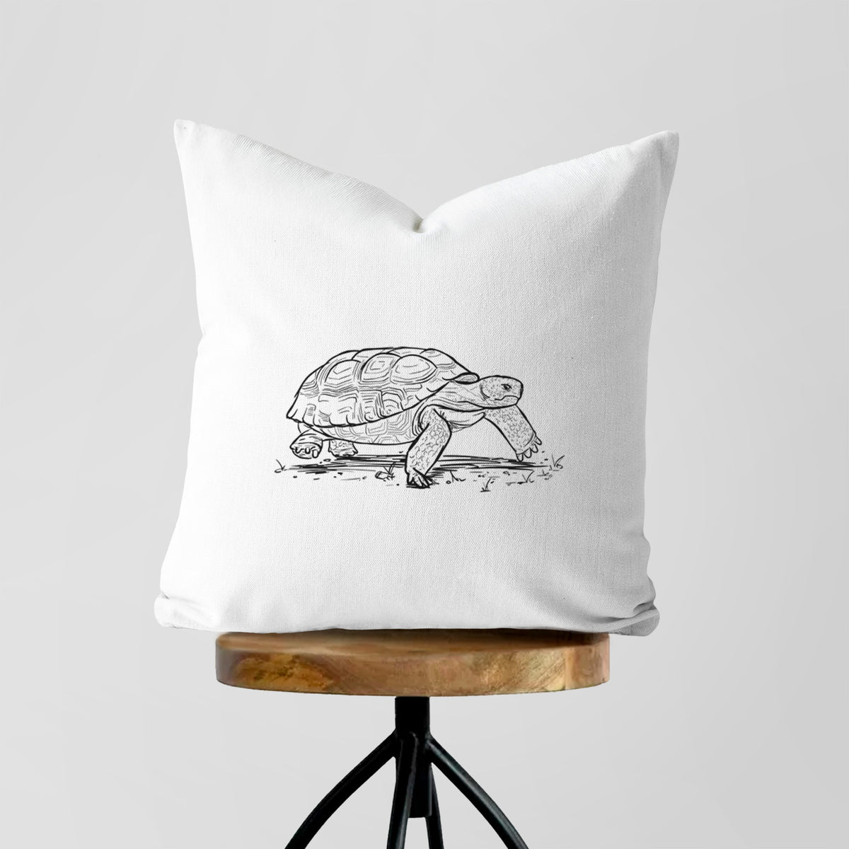 White pillow cover printed with a detailed black ink illustration of a gopher tortoise walking, on a wooden stool.