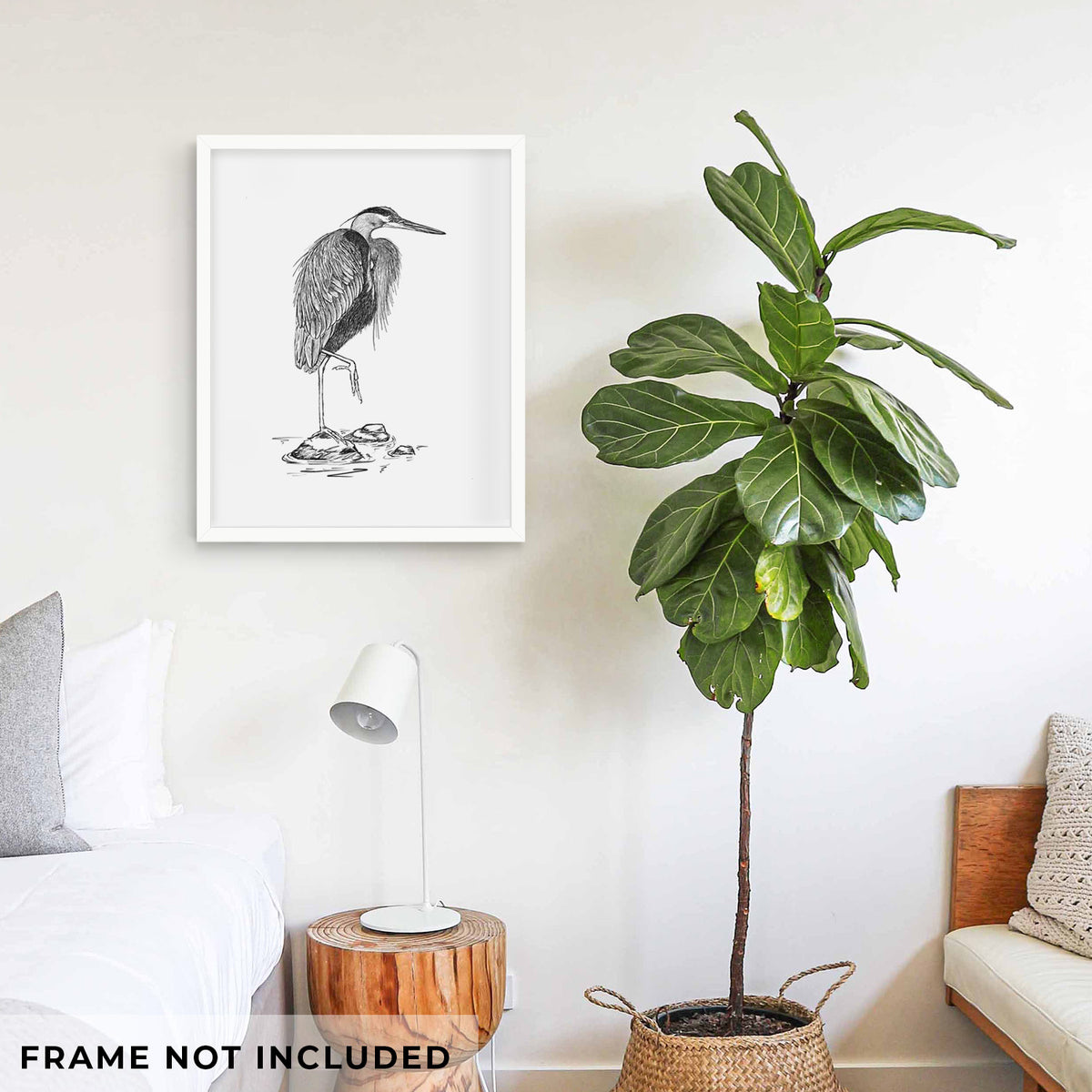Art print of a detailed black ink illustration of a great blue heron standing on rocks, shown framed on a living room wall beside a potted plant. Frame not included.