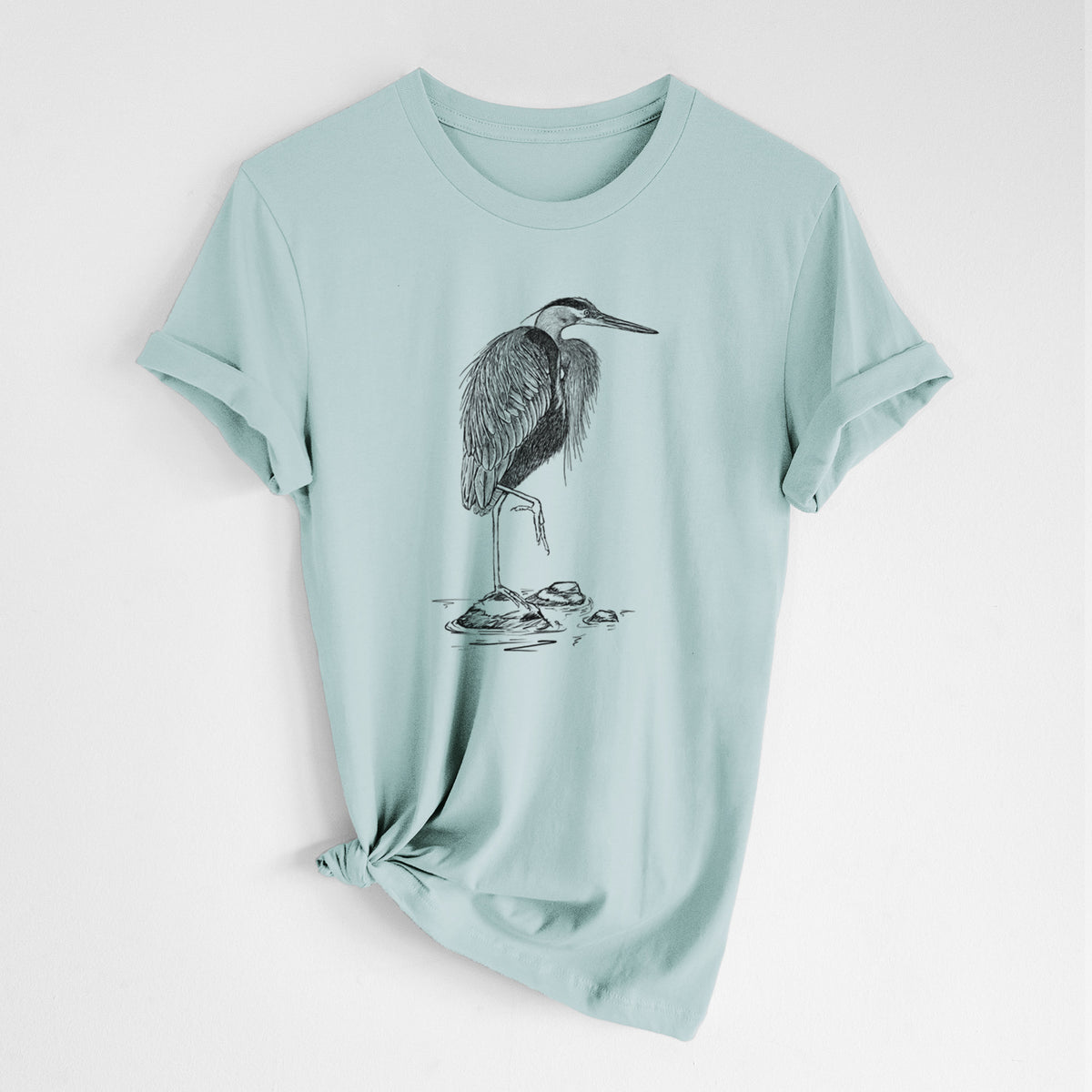 Dusty blue lightweight cotton t-shirt laid flat with the sleeves rolled, printed with a detailed black ink illustration of a great blue heron standing on rocks.