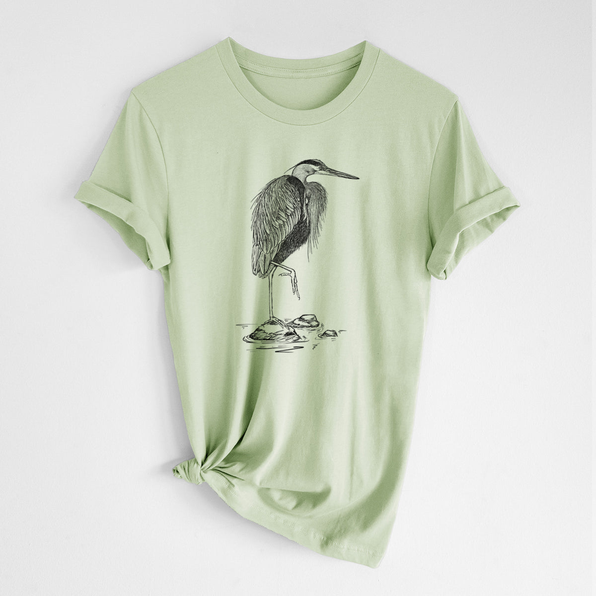 Spring green lightweight cotton t-shirt laid flat with the sleeves rolled, printed with a detailed black ink illustration of a great blue heron standing on rocks.