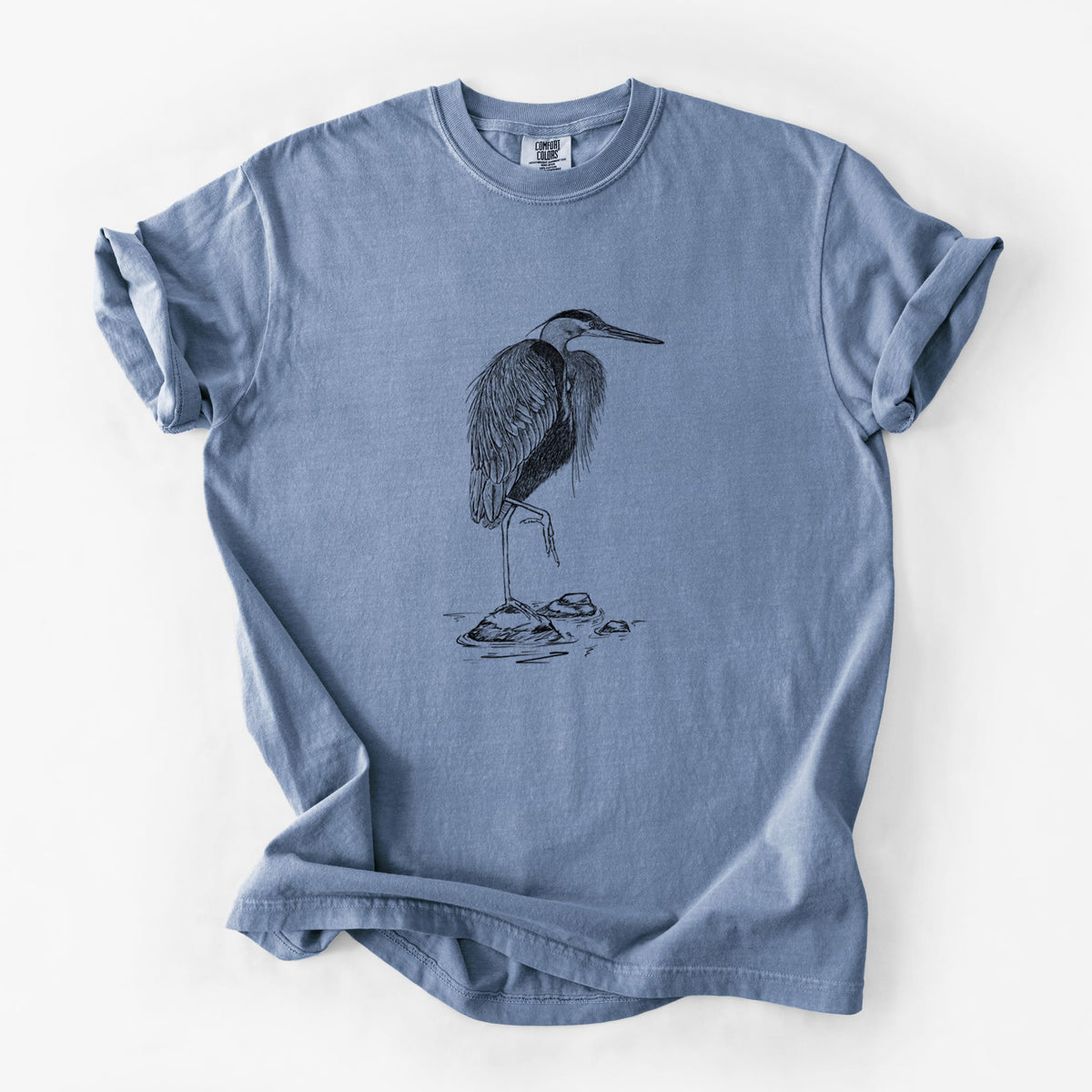 Blue jean Comfort Colors t-shirt laid flat, printed with a detailed black ink illustration of a great blue heron standing on rocks.
