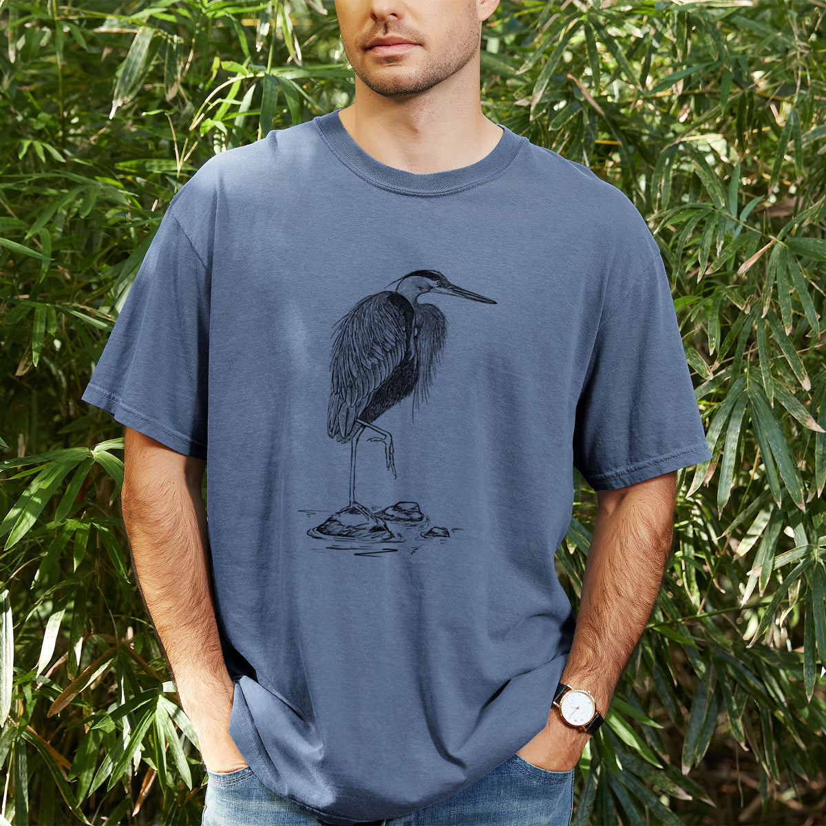 Man wearing a Comfort Colors t-shirt printed with a detailed black ink illustration of a great blue heron standing on rocks, outdoors.