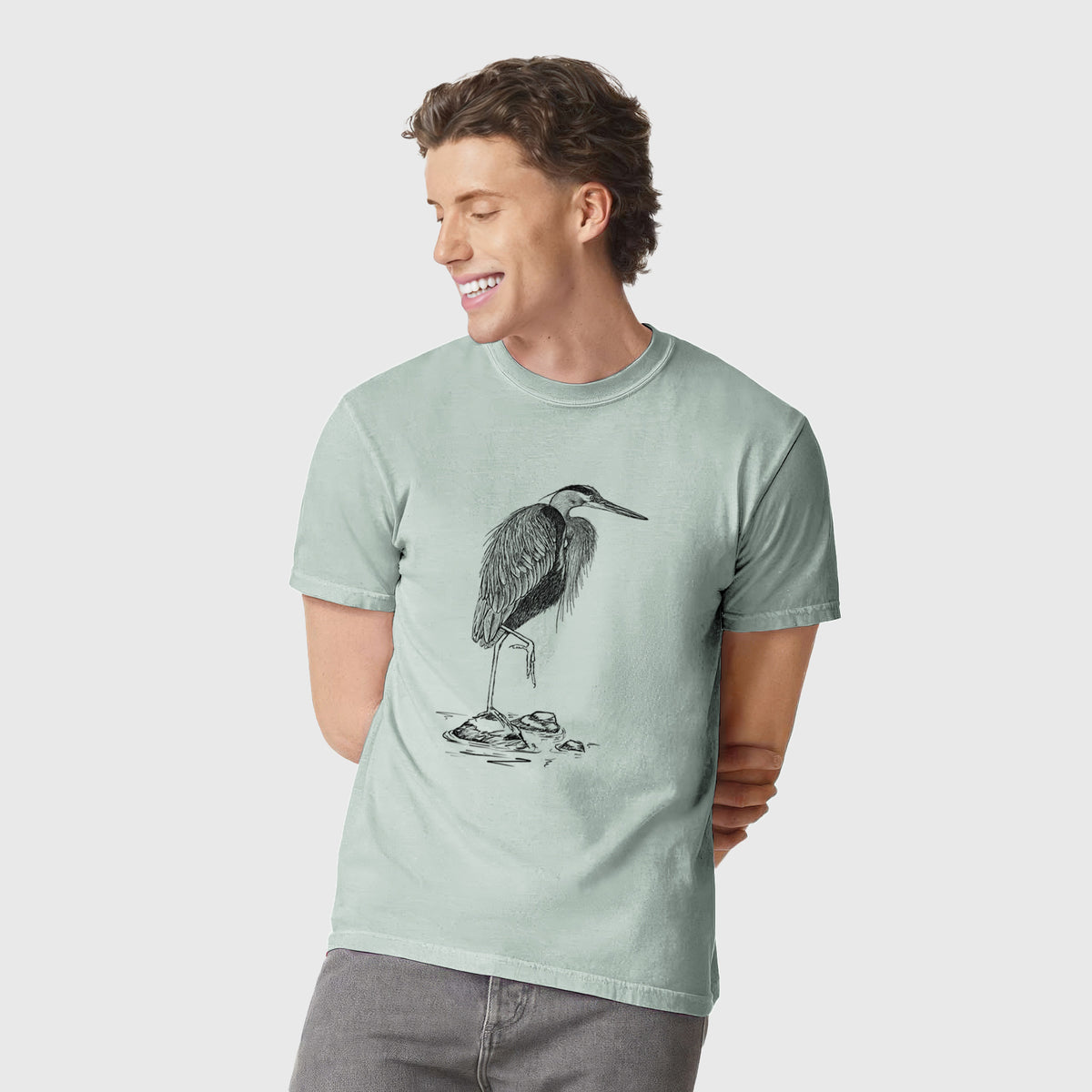 Man wearing a Comfort Colors t-shirt printed with a detailed black ink illustration of a great blue heron standing on rocks.