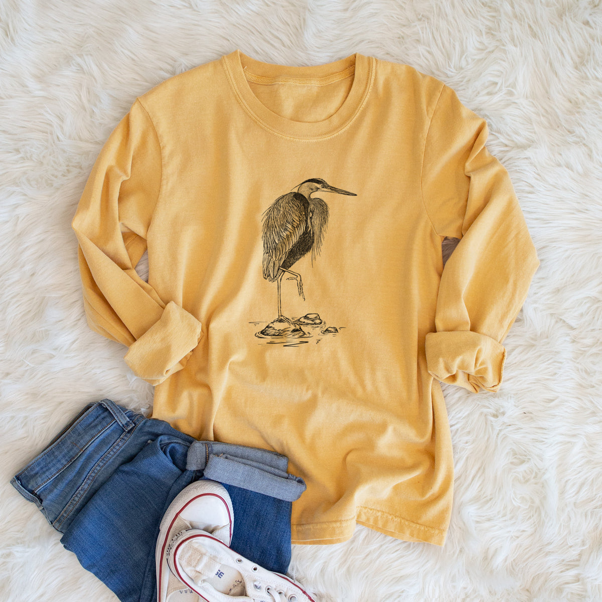 Golden wheat yellow Comfort Colors long sleeve shirt printed with a detailed black ink illustration of a great blue heron standing on rocks, shown in a lifestyle photo.