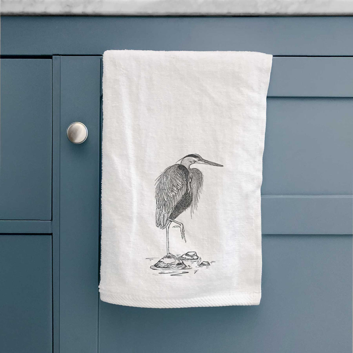 White hand towel printed with a detailed black ink illustration of a great blue heron standing on rocks, hanging on a cabinet door.