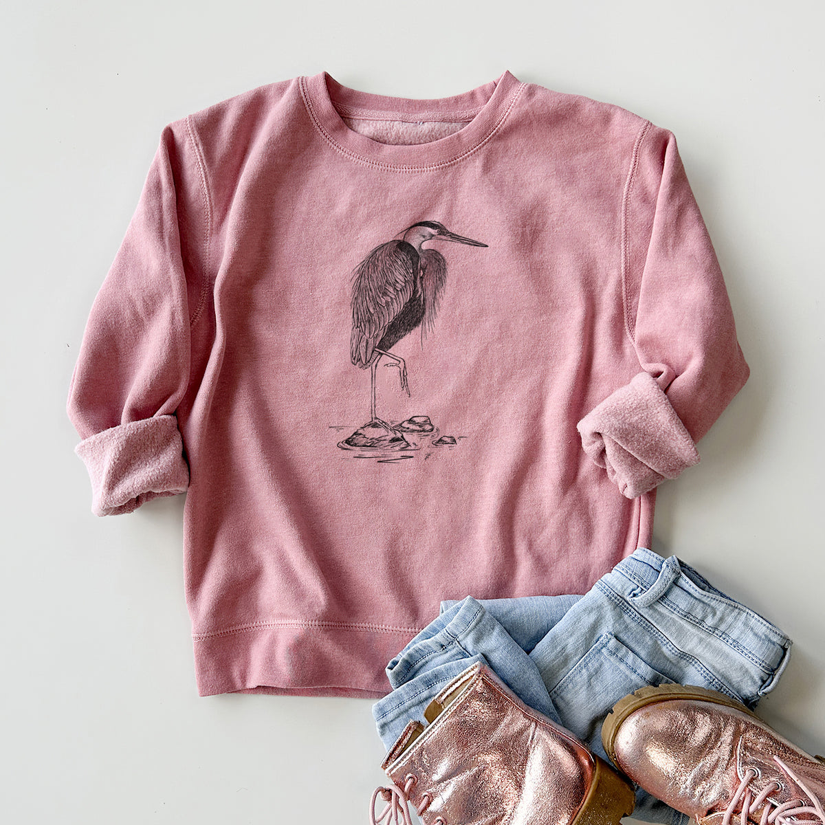 Youth crewneck sweatshirt printed with a detailed black ink illustration of a great blue heron standing on rocks, styled flat with jeans and boots.