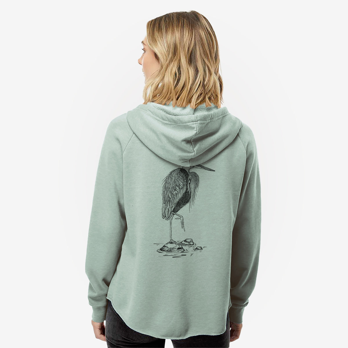 Woman wearing a sage green women's Cali Wave zip-up sweatshirt printed with a detailed black ink illustration of a great blue heron standing on rocks, seen from behind.