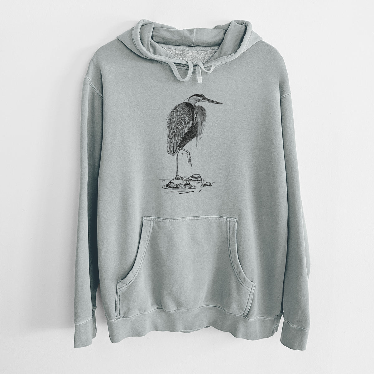 Sage green pigment dyed hoodie laid flat, printed with a detailed black ink illustration of a great blue heron standing on rocks.