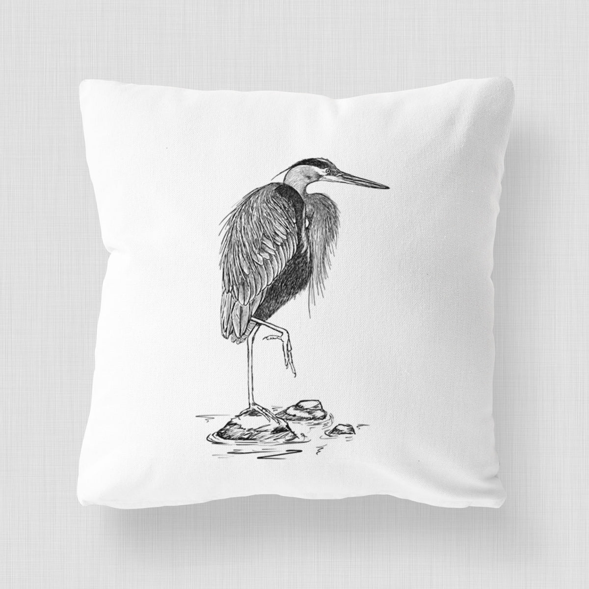 White pillow cover printed with a detailed black ink illustration of a great blue heron standing on rocks.