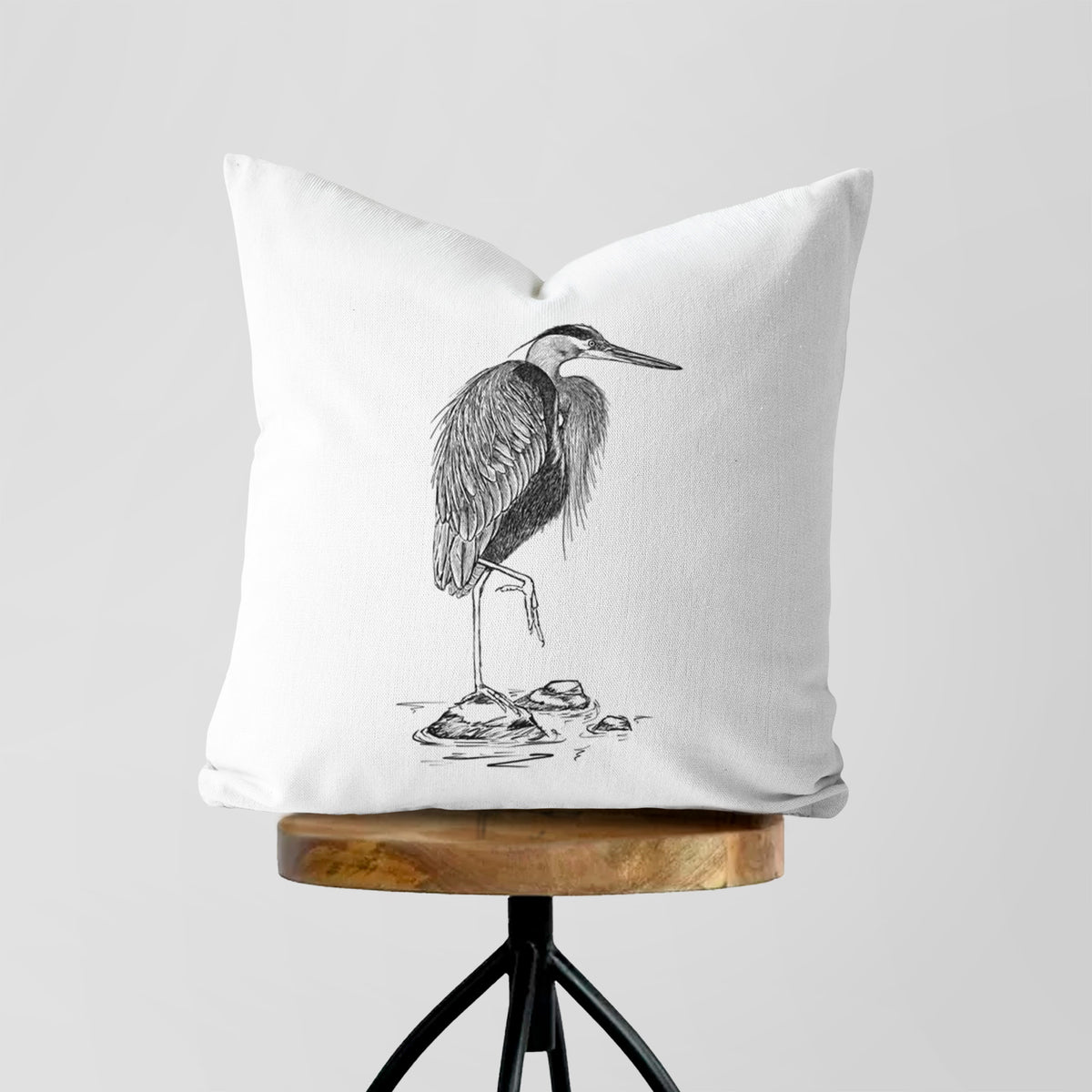 White pillow cover printed with a detailed black ink illustration of a great blue heron standing on rocks, on a wooden stool.