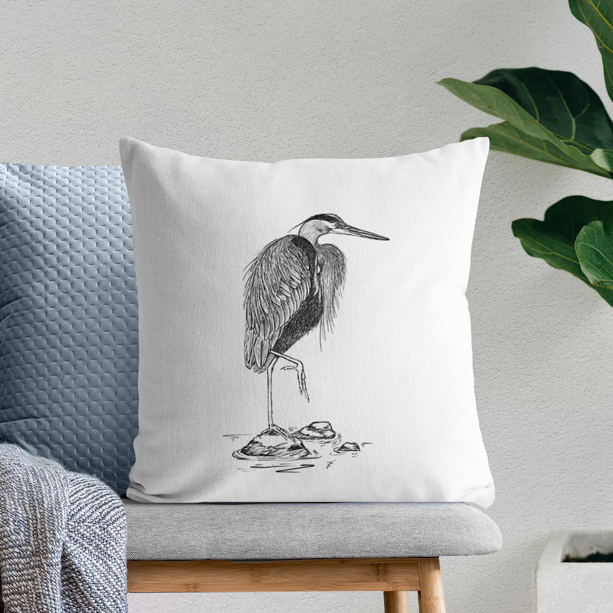 White pillow cover printed with a detailed black ink illustration of a great blue heron standing on rocks, on a bench beside plants.