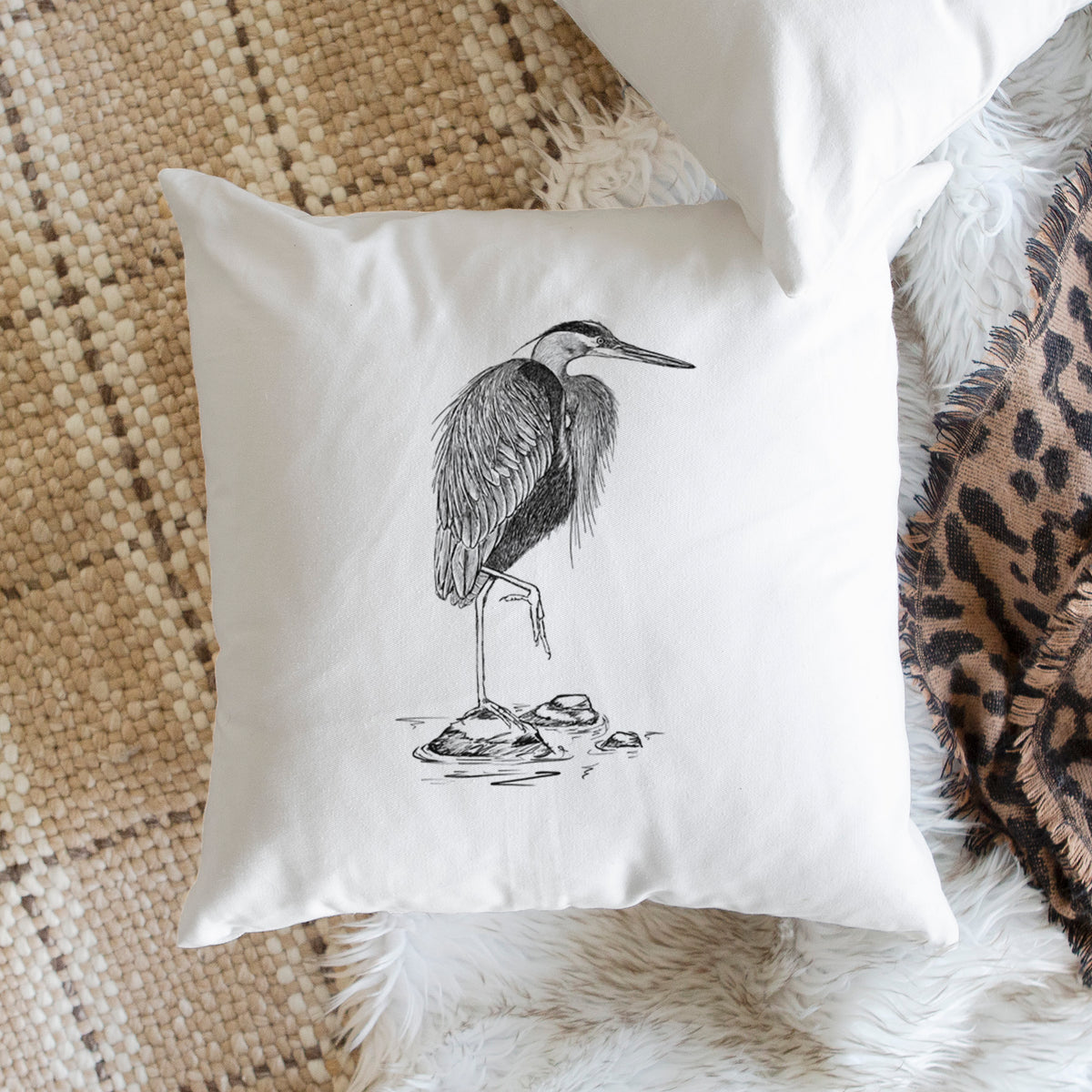 White pillow cover printed with a detailed black ink illustration of a great blue heron standing on rocks, on a woven chair.