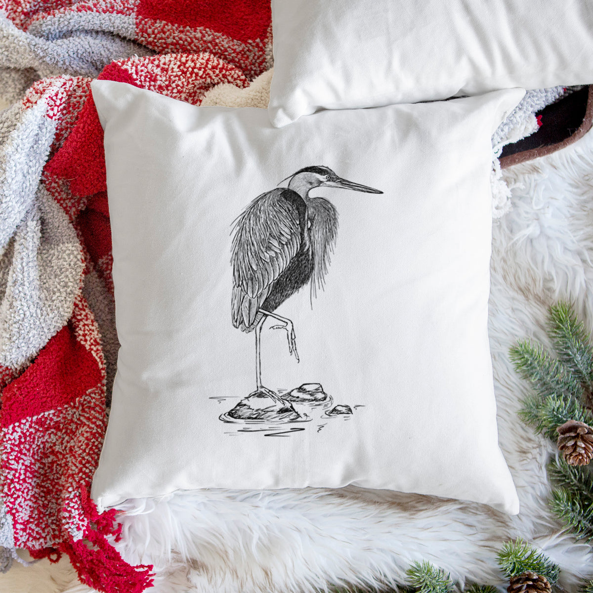 White pillow cover printed with a detailed black ink illustration of a great blue heron standing on rocks, on a woven chair.