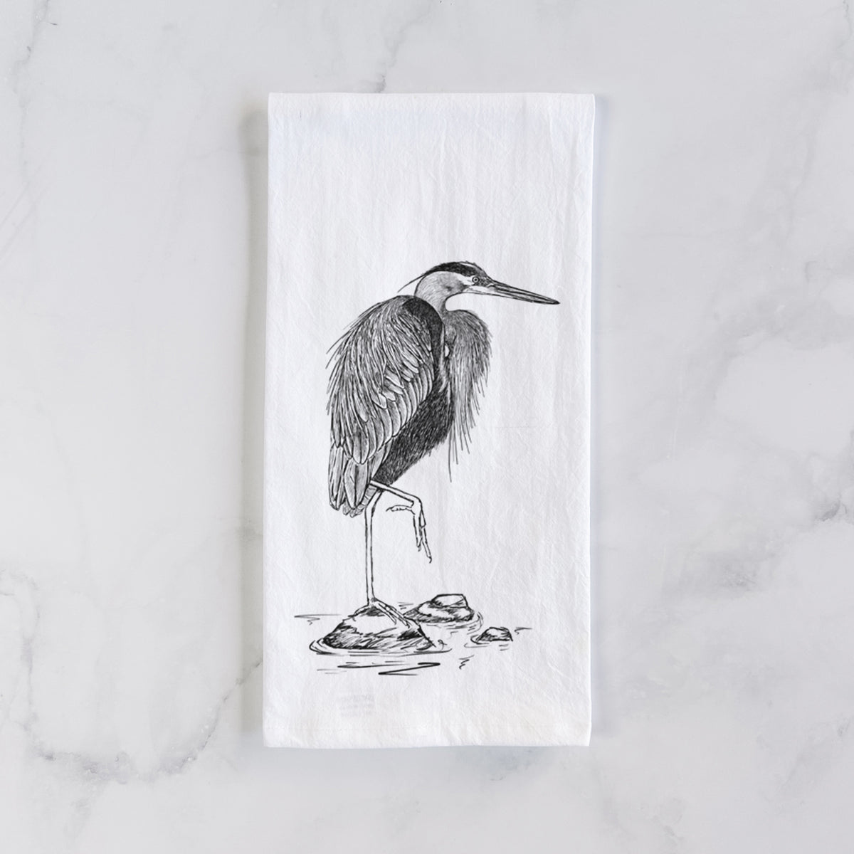 White cotton tea towel laid flat on marble, printed with a detailed black ink illustration of a great blue heron standing on rocks.