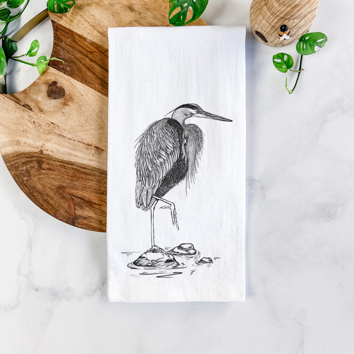 White cotton tea towel printed with a detailed black ink illustration of a great blue heron standing on rocks, on a marble counter beside a round wooden cutting board and greenery.