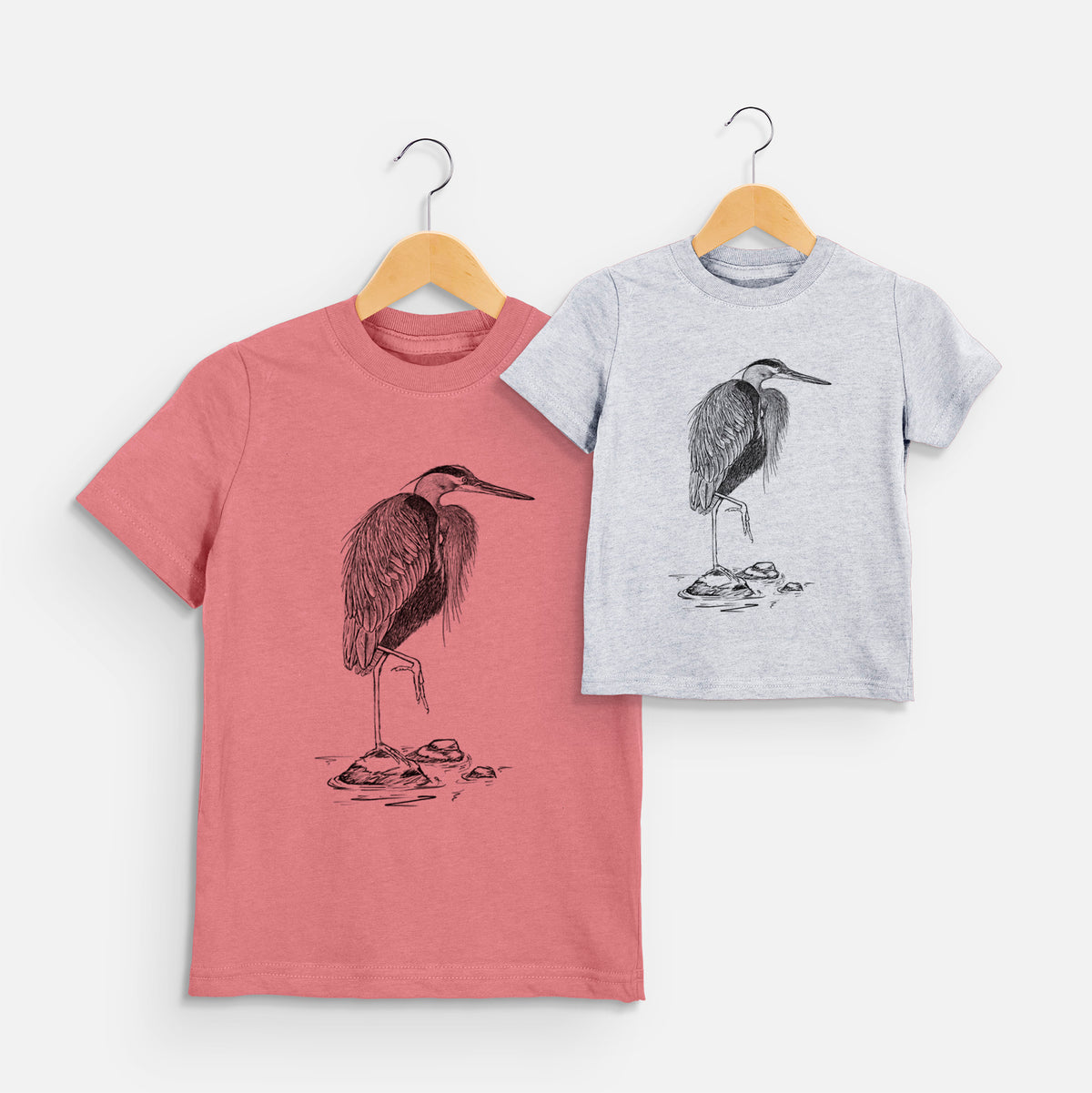 Two youth t-shirts in different colors on hangers, printed with a detailed black ink illustration of a great blue heron standing on rocks.