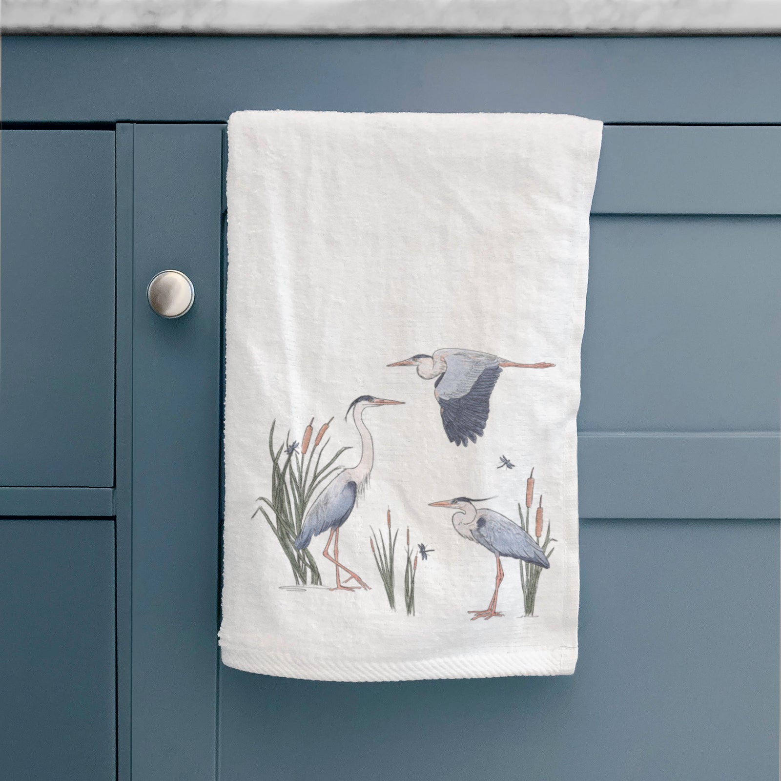 White hand towel printed with a full-color illustration of three great blue herons among cattails, one in flight.