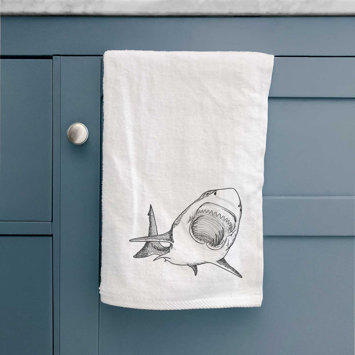 White hand towel printed with a black ink illustration of a great white shark swimming toward the viewer with its mouth open, hanging on a cabinet door.
