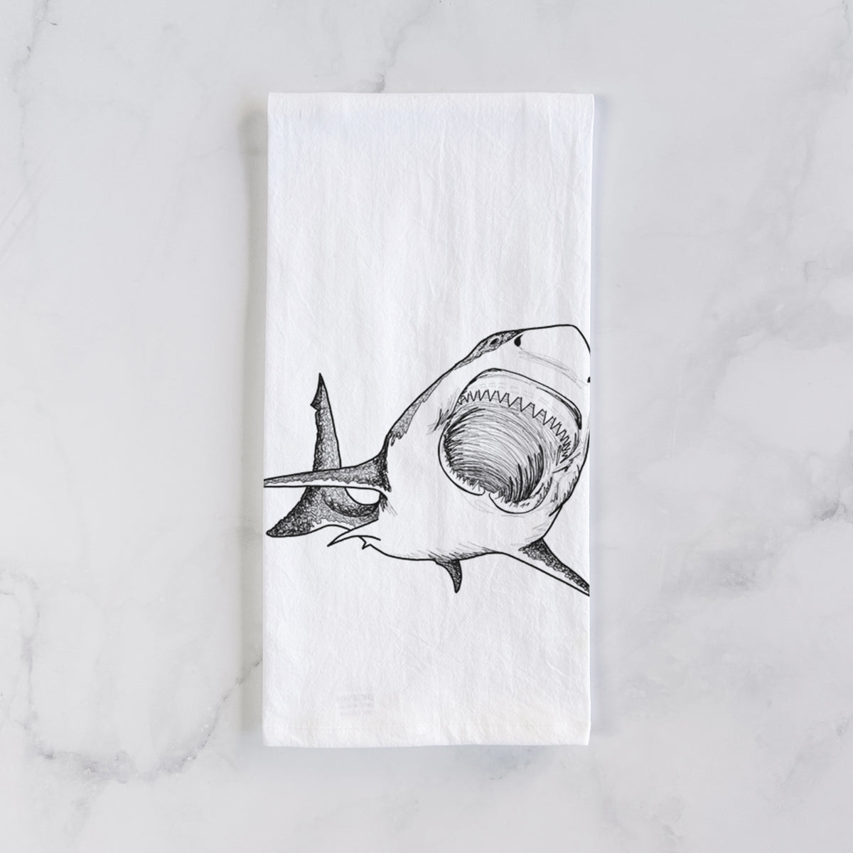 White cotton tea towel laid flat on marble, printed with a black ink illustration of a great white shark swimming toward the viewer with its mouth open.