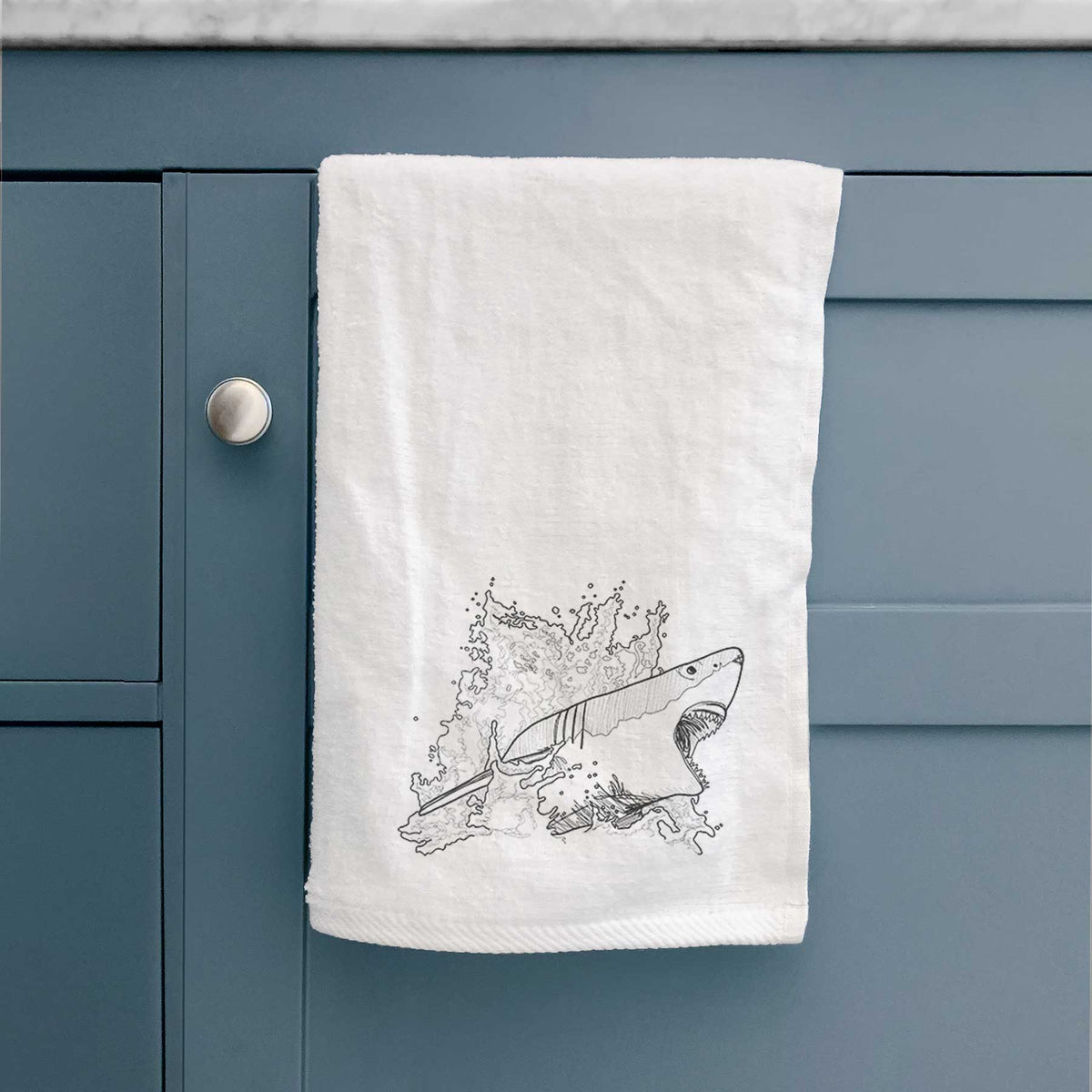 White hand towel printed with a black ink illustration of a great white shark breaking through a splash of water, hanging on a cabinet door.