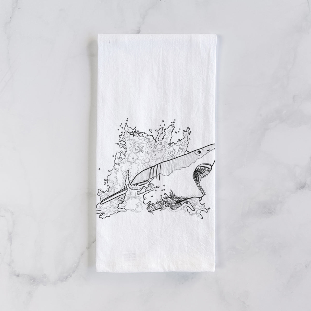 White cotton tea towel laid flat on marble, printed with a black ink illustration of a great white shark breaking through a splash of water.
