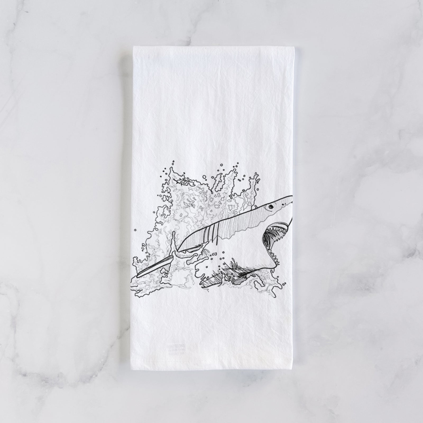 White cotton tea towel printed with a black ink illustration of a great white shark breaking through a splash of water, on a marble counter beside a round wooden cutting board and greenery.