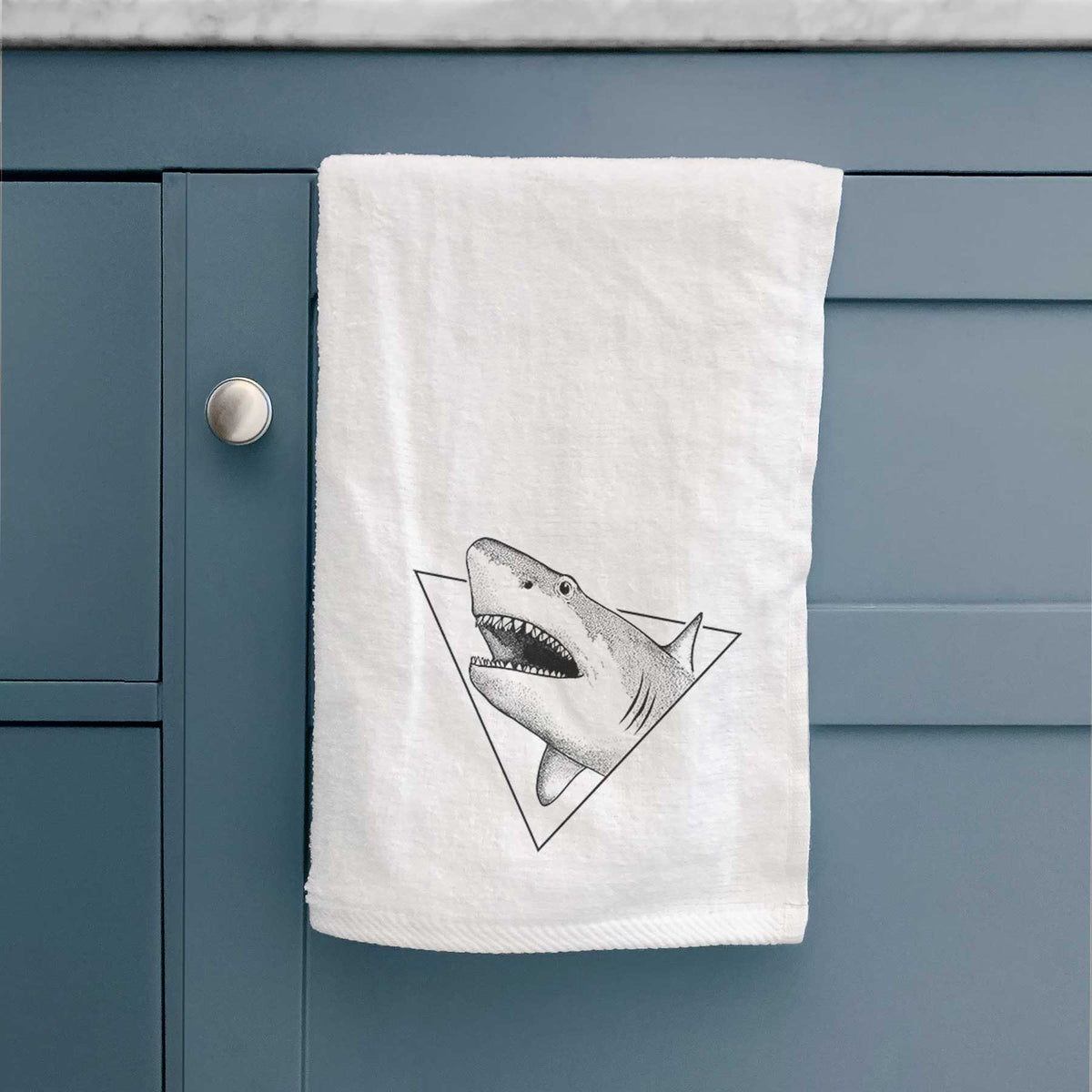 White hand towel printed with a black ink great white shark bursting out of a triangle outline with its mouth open, hanging on a cabinet door.
