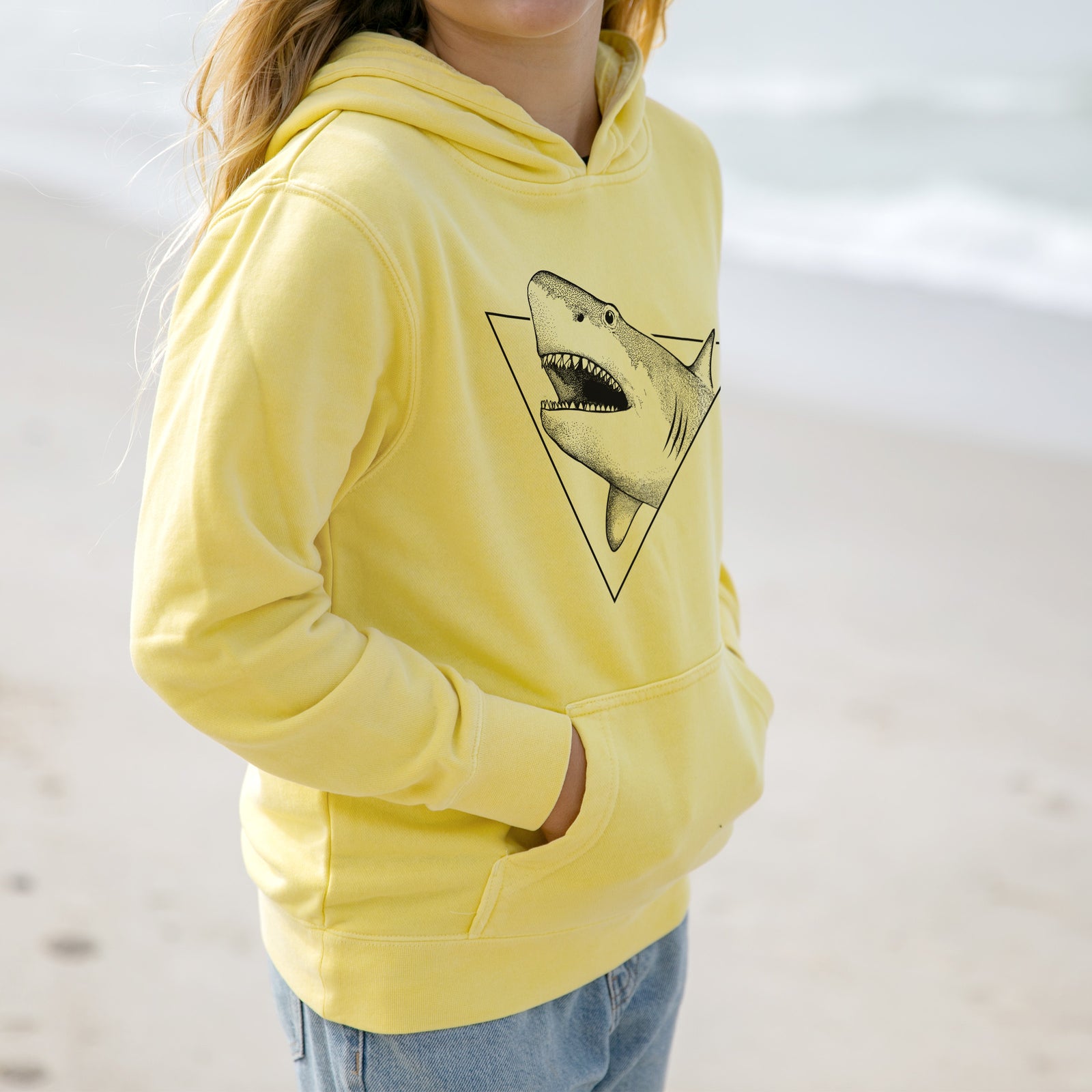 Boy wearing a youth pigment dyed hoodie printed with a black ink great white shark bursting out of a triangle outline with its mouth open.