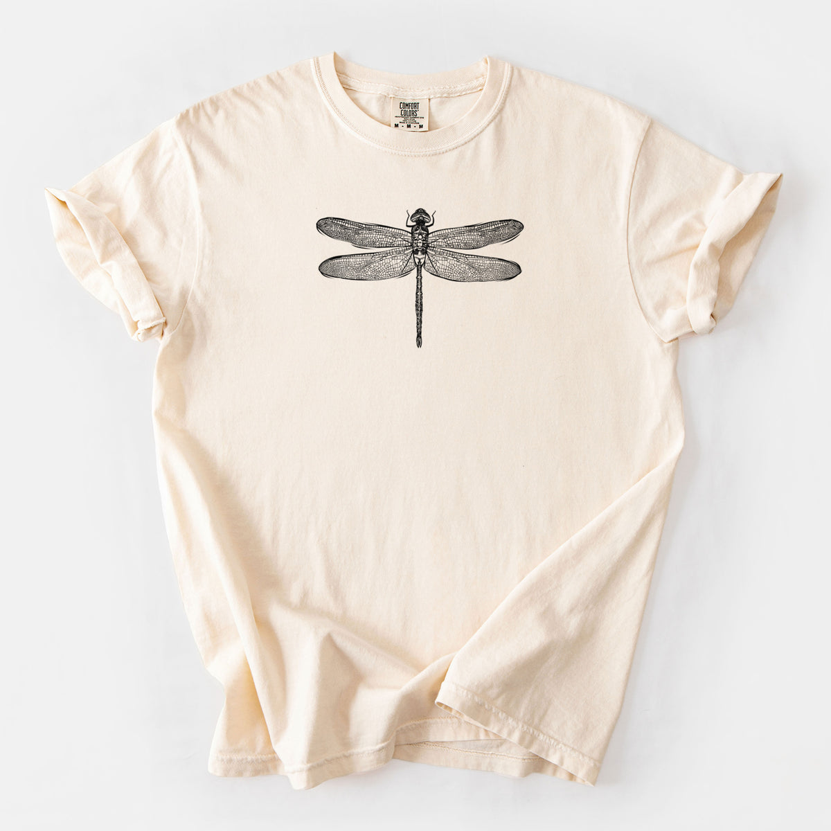 Ivory Comfort Colors t-shirt laid flat, printed with a detailed black ink illustration of a green darner dragonfly with its wings spread, seen from above.