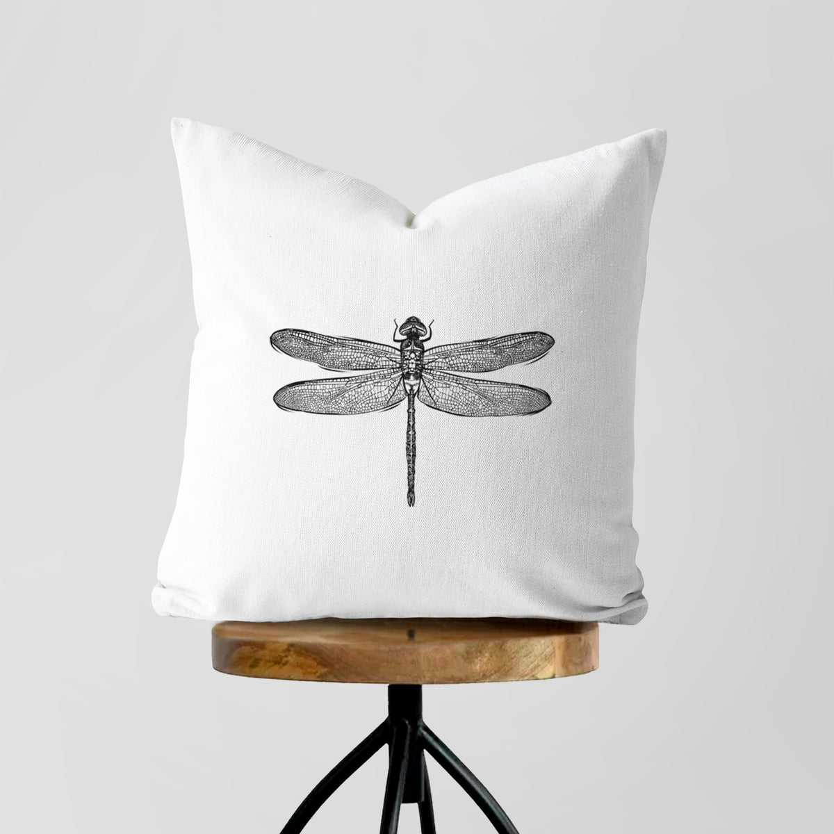 White pillow cover printed with a detailed black ink illustration of a green darner dragonfly with its wings spread, seen from above, on a wooden stool.