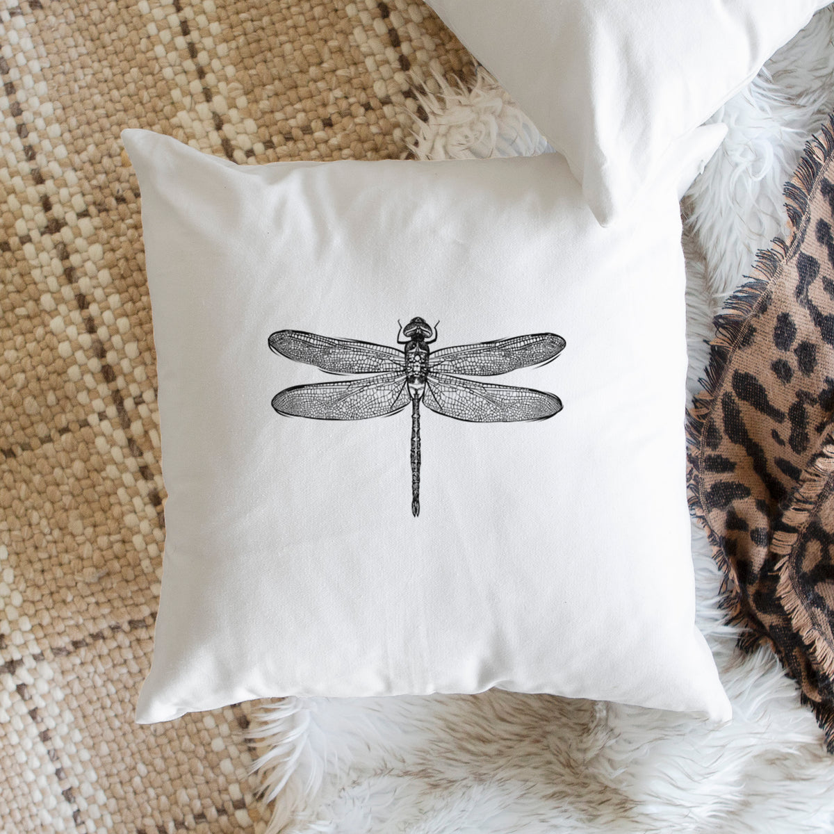 White pillow cover printed with a detailed black ink illustration of a green darner dragonfly with its wings spread, seen from above, on a woven chair.
