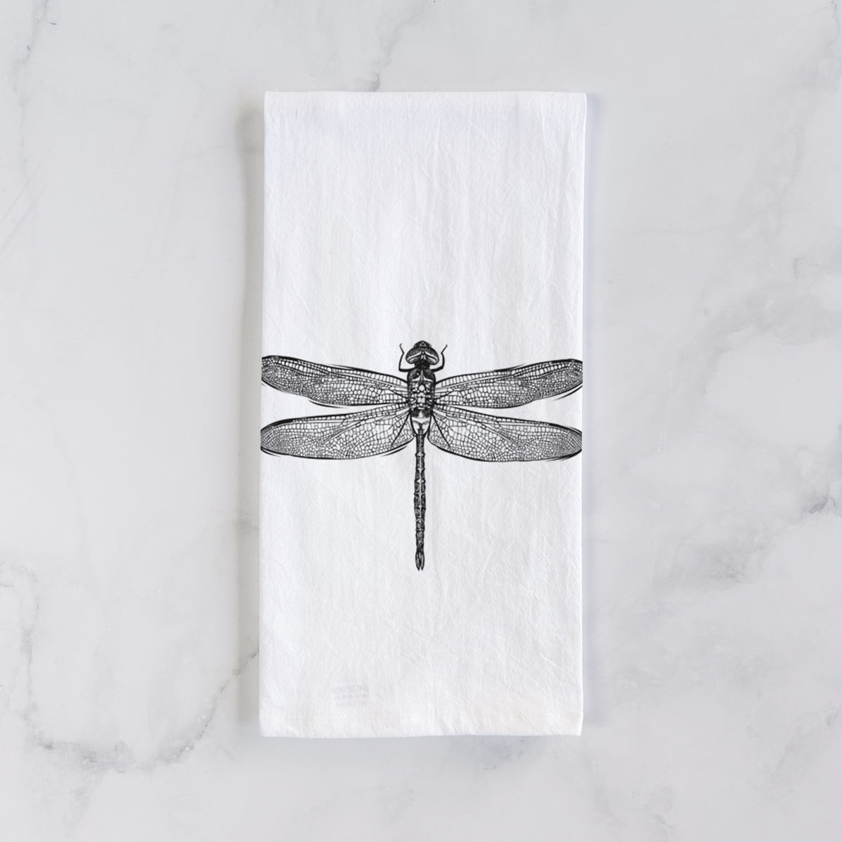 White cotton tea towel laid flat on marble, printed with a detailed black ink illustration of a green darner dragonfly with its wings spread, seen from above.