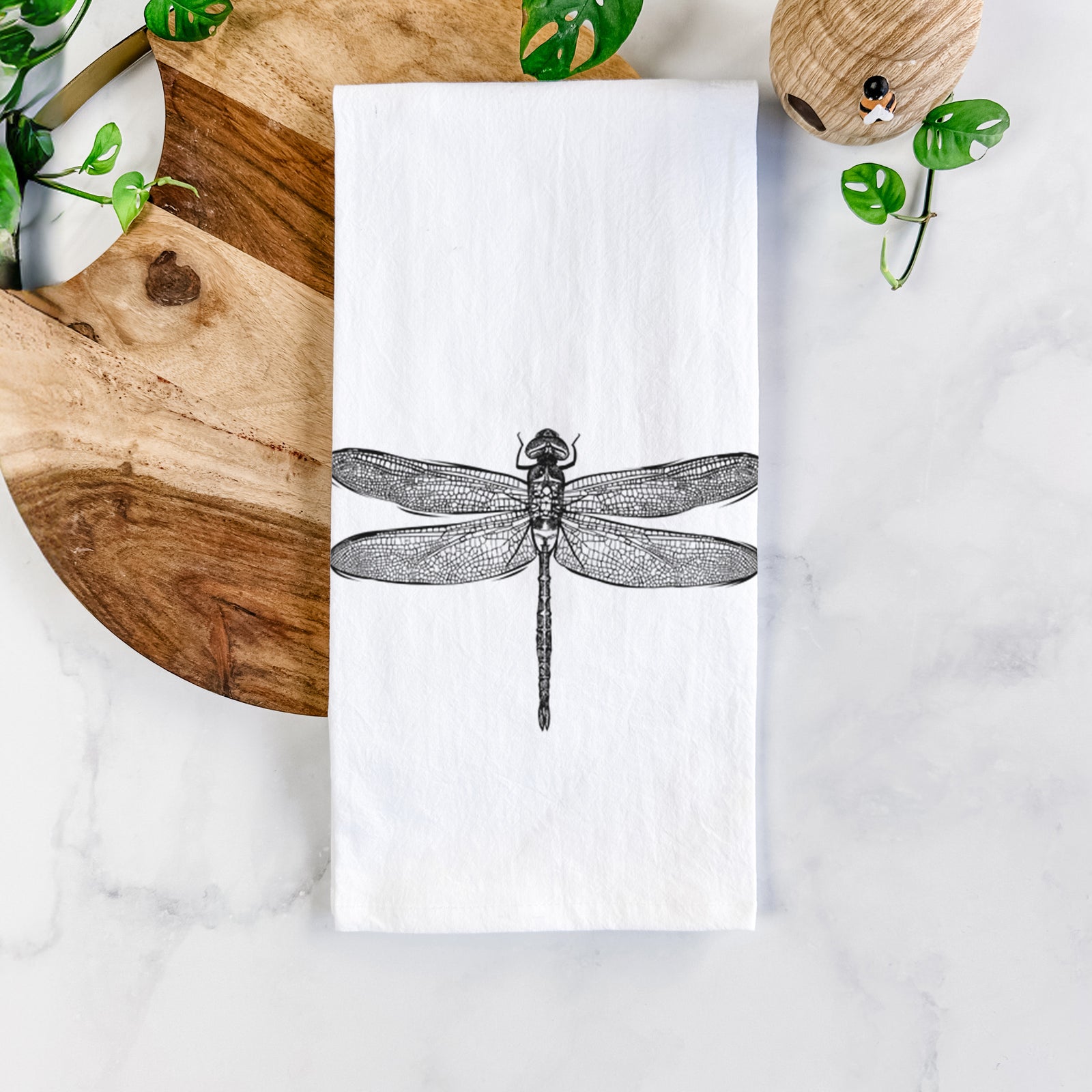 White cotton tea towel printed with a detailed black ink illustration of a green darner dragonfly with its wings spread, seen from above, on a marble counter beside a round wooden cutting board and greenery.