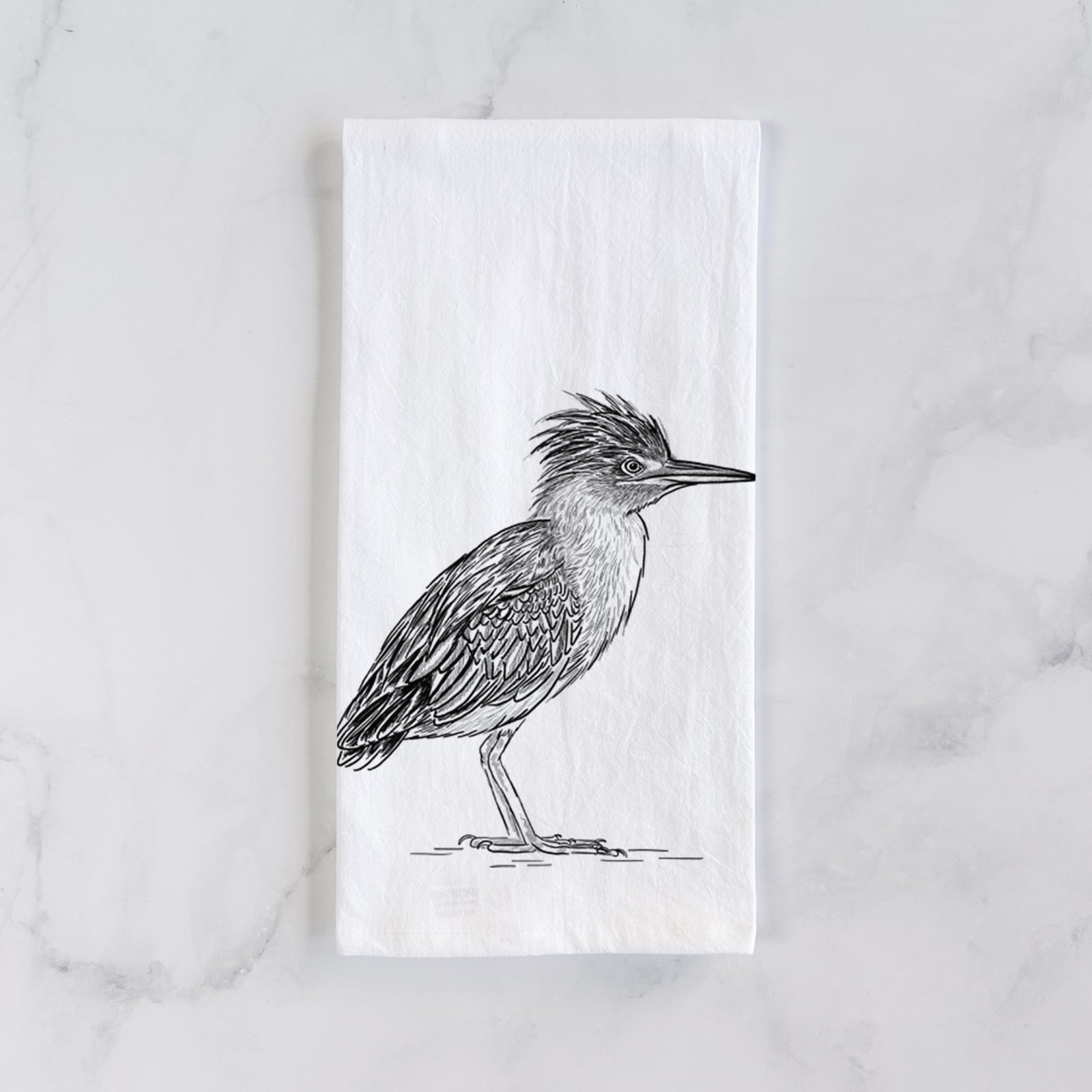 White cotton tea towel printed with a detailed black ink illustration of a green heron standing with its crest raised, on a marble counter beside a round wooden cutting board and greenery.