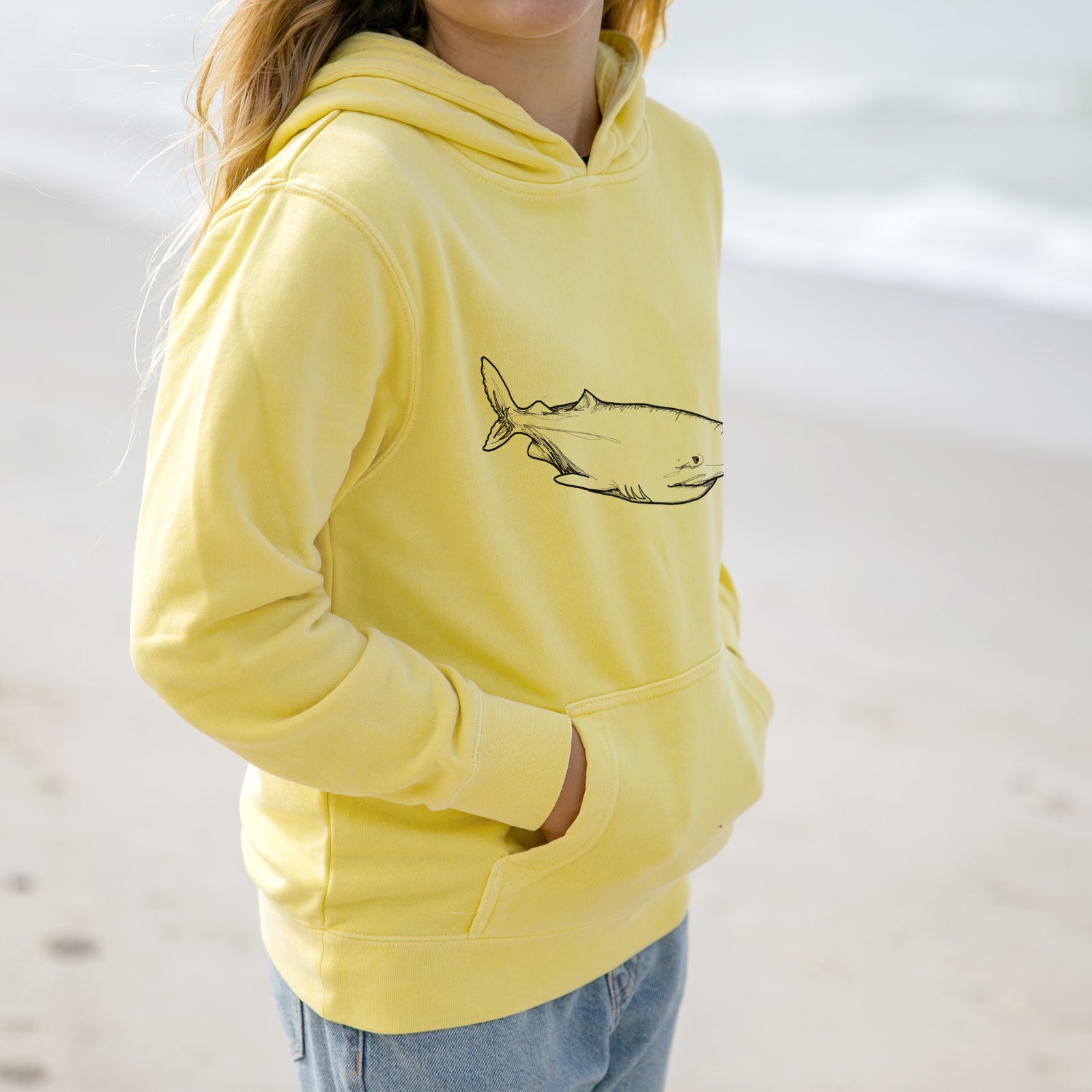 Boy wearing a youth pigment dyed hoodie printed with a black ink illustration of a Greenland shark swimming.
