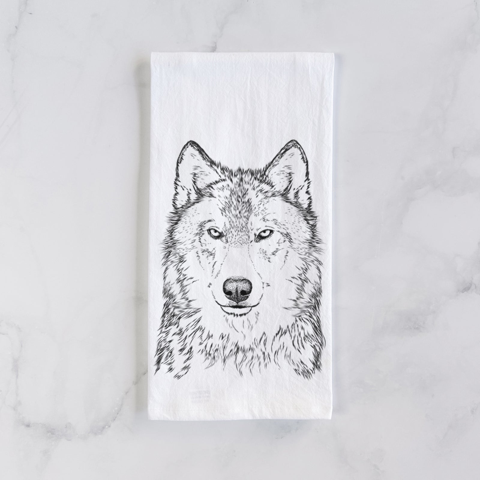White cotton tea towel printed with a detailed black ink portrait of a grey wolf facing forward, on a marble counter beside a round wooden cutting board and greenery.