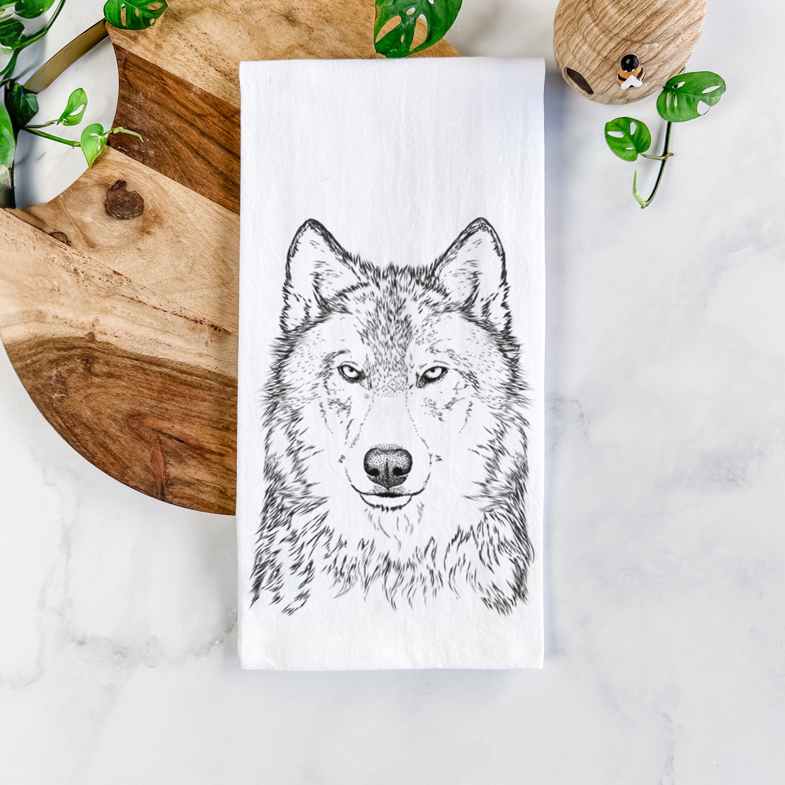 White cotton tea towel printed with a detailed black ink portrait of a grey wolf facing forward, on a marble counter beside a round wooden cutting board and greenery.