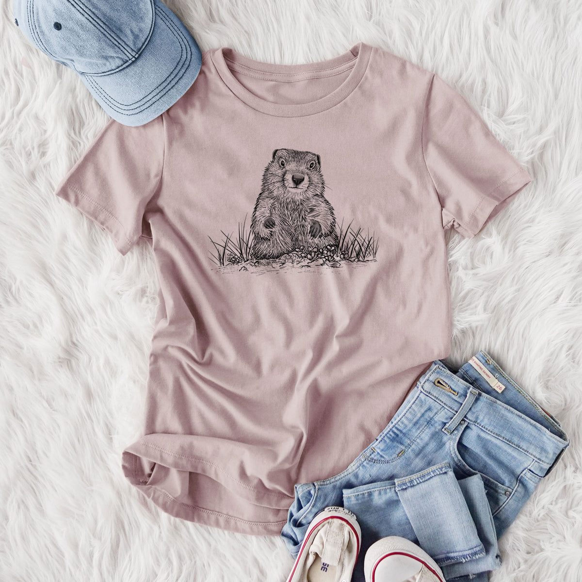 Women's relaxed fit cotton t-shirt printed with a black ink illustration of a groundhog peeking out of its burrow among grass, styled flat on a white faux fur rug with jeans.