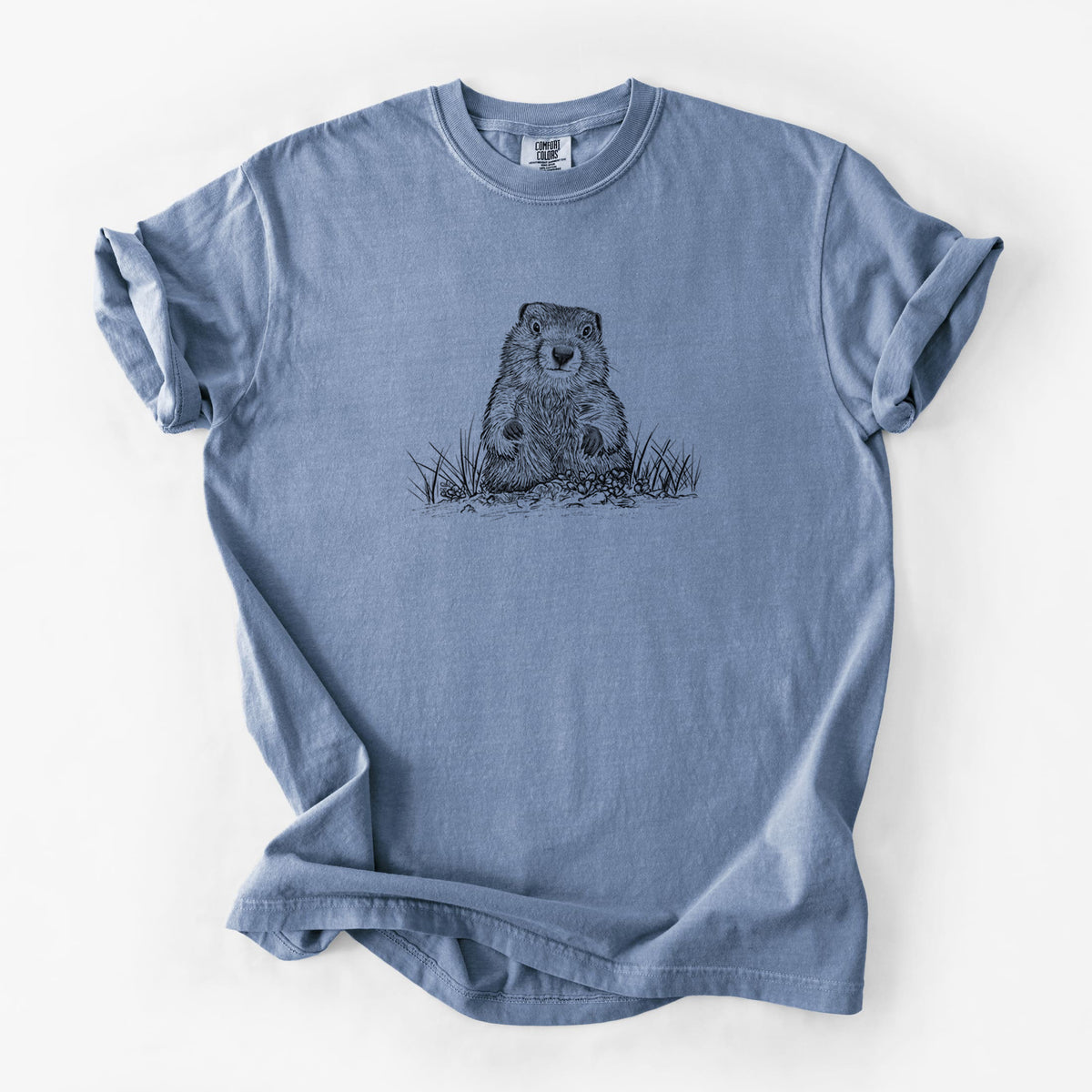 Blue jean Comfort Colors t-shirt laid flat, printed with a black ink illustration of a groundhog peeking out of its burrow among grass.
