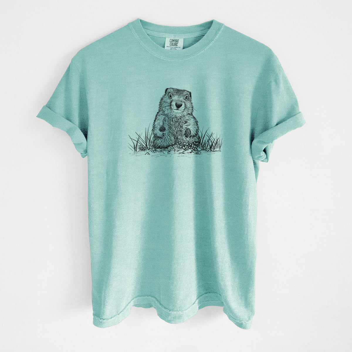 Chalky mint Comfort Colors t-shirt laid flat, printed with a black ink illustration of a groundhog peeking out of its burrow among grass.